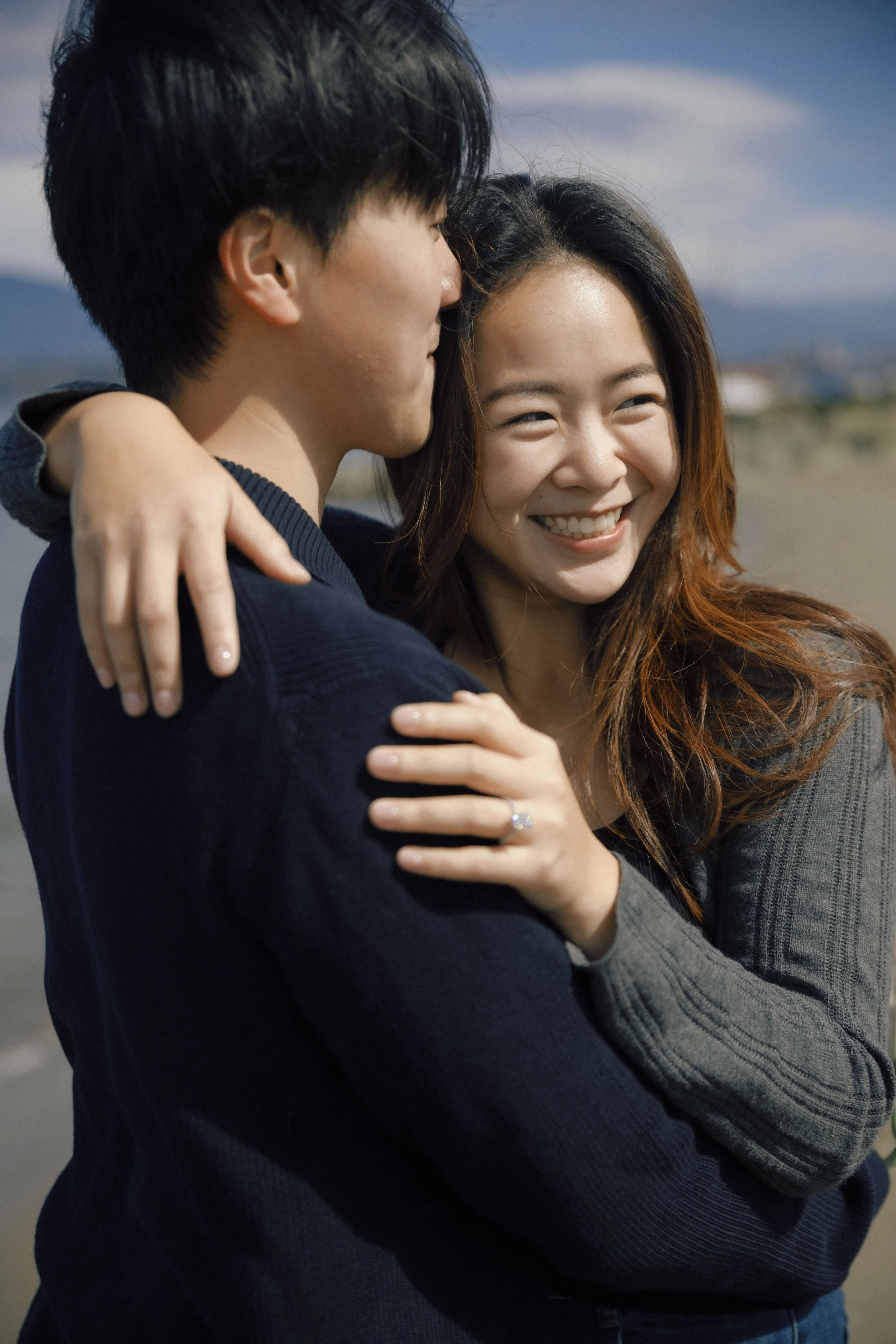 A young woman smiling and hugging a young man outside on a cloudy day with mountains and sky in the background.