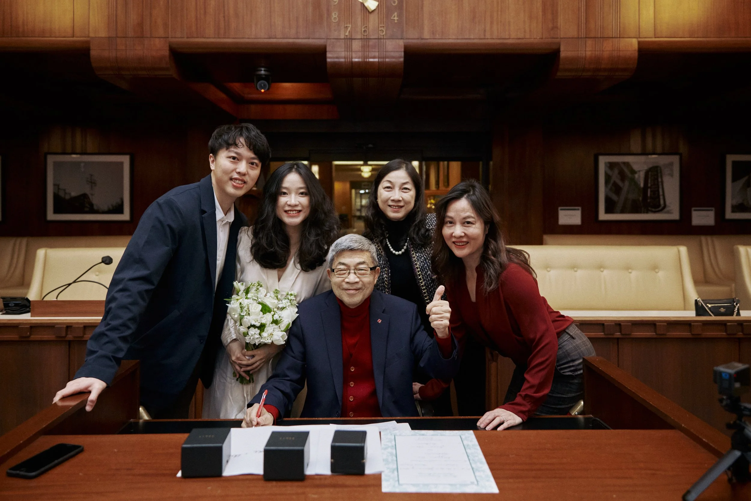 A group of five people in a formal setting, with one elderly man signing a document, surrounded by four younger individuals, one holding a bouquet of flowers.
