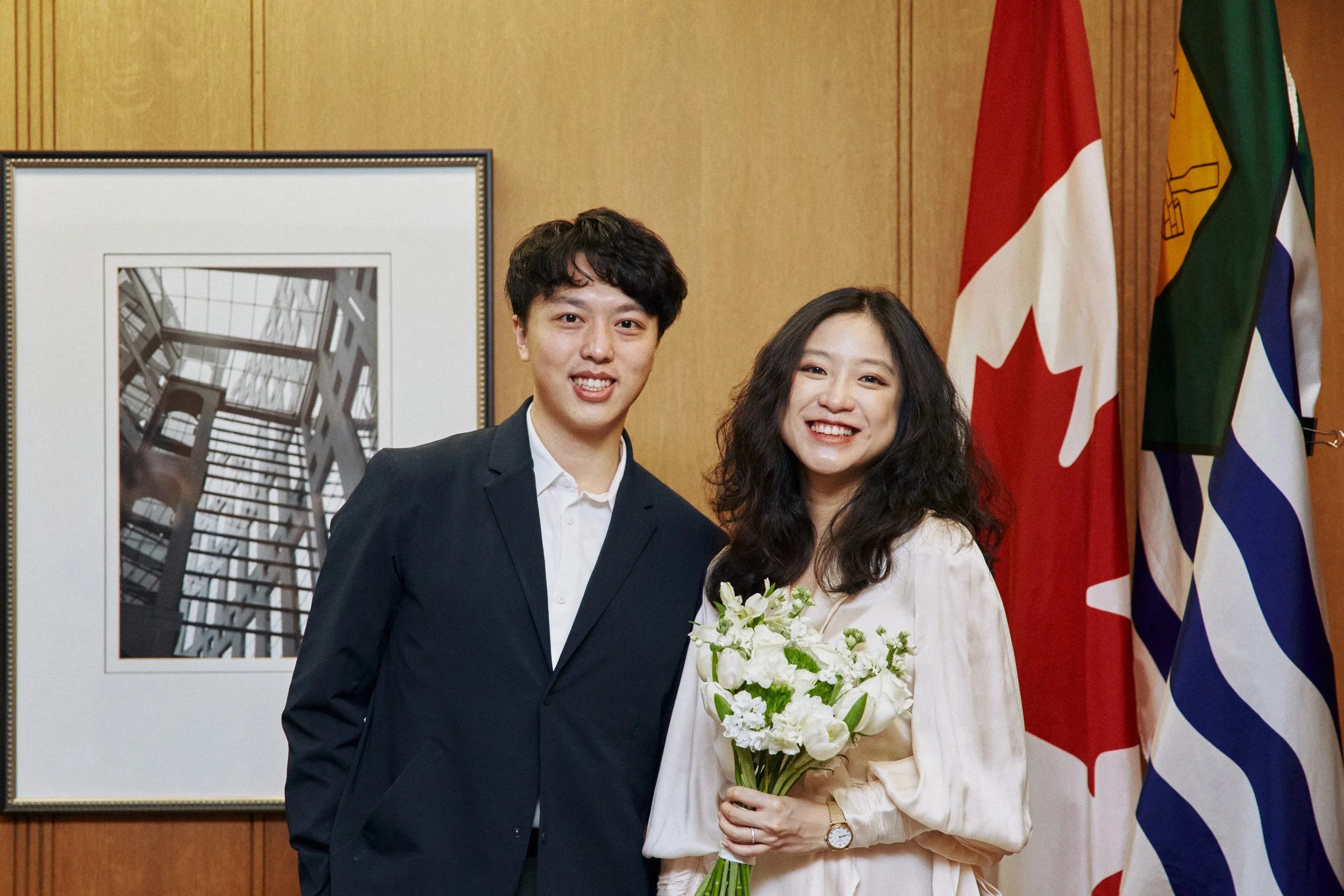 A young man in a dark suit and a young woman in a cream-colored dress are standing together, smiling at the camera. The woman is holding a bouquet of white flowers. They are indoors, standing in front of a wooden wall with flags and a picture of a bu