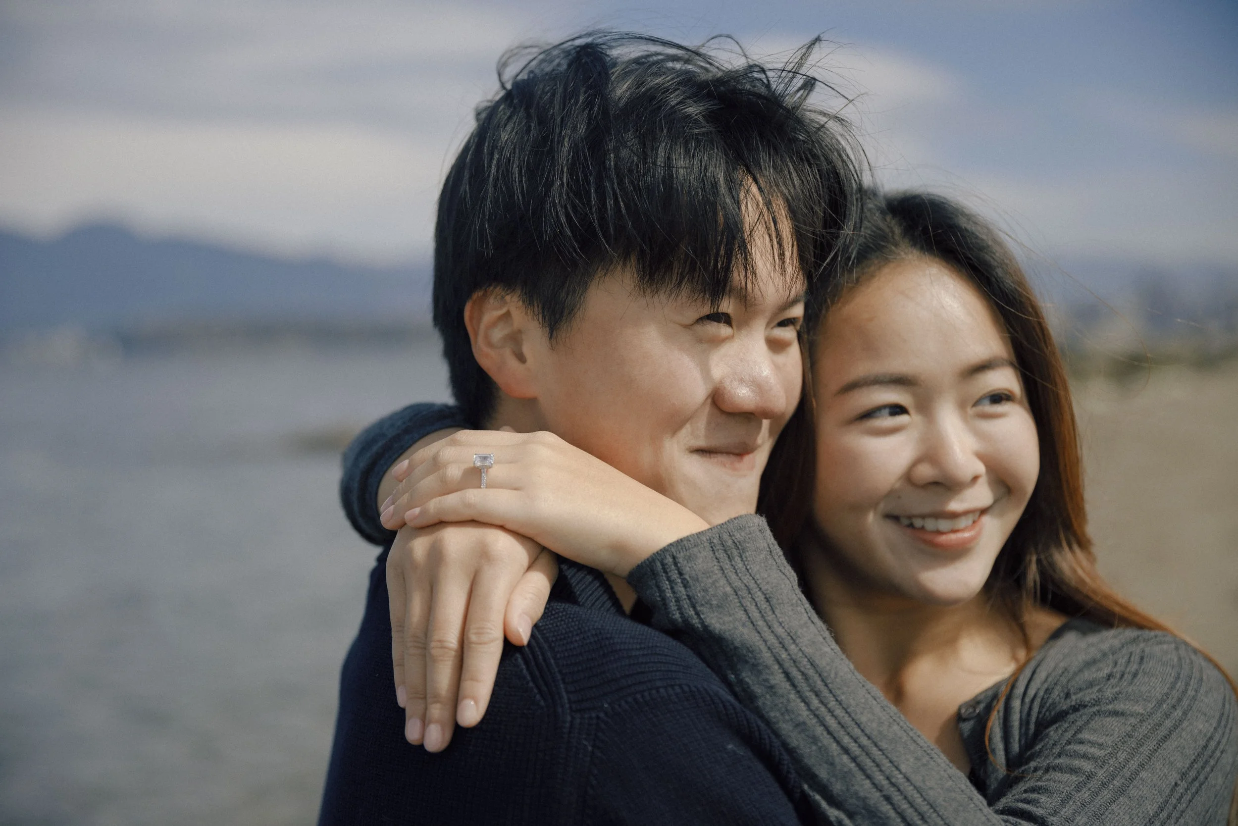 A young man and woman hugging outdoors with a body of water and mountains in the background, smiling.