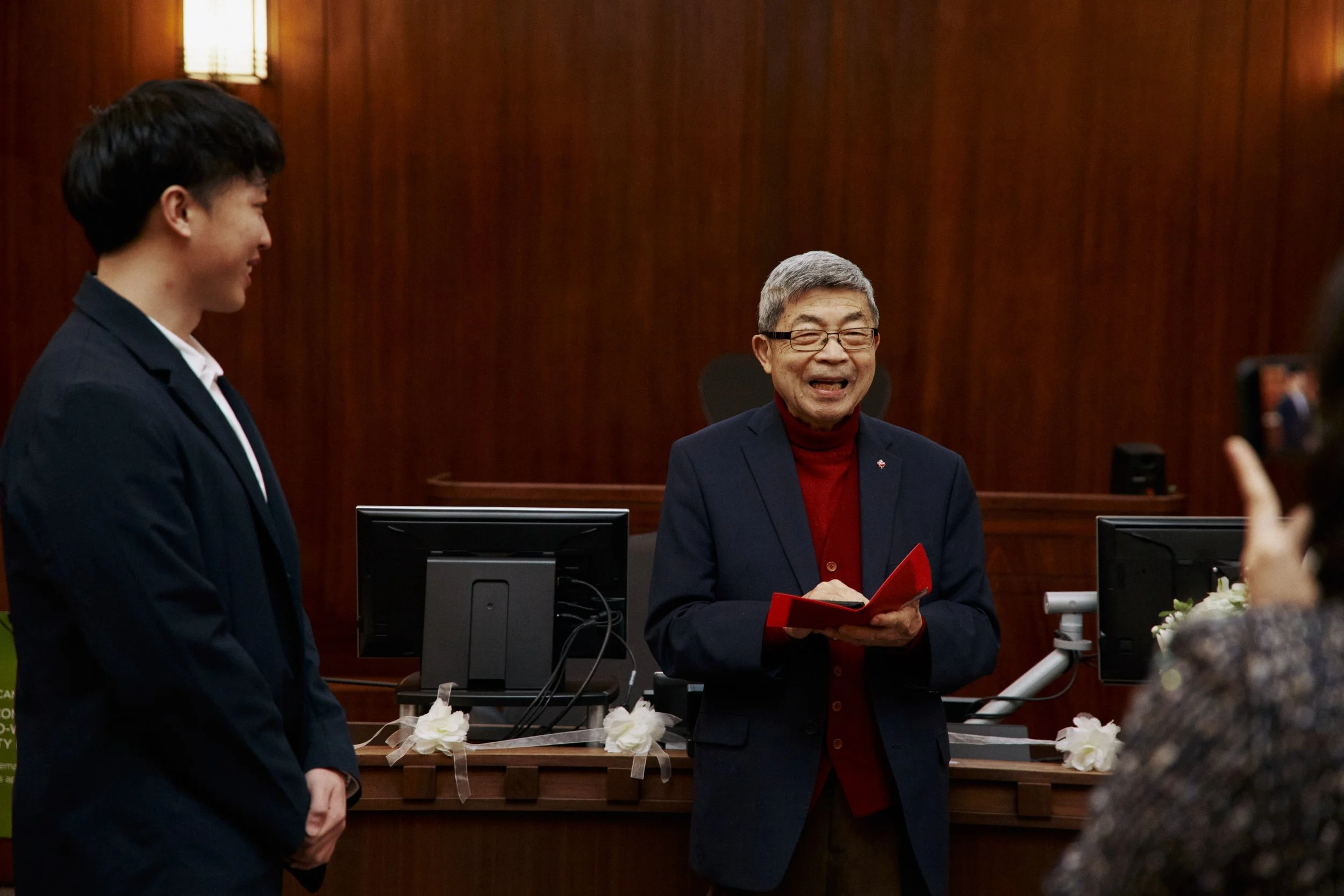 An older man with gray hair and glasses, wearing a red turtleneck and a dark blazer, is holding a red document folder and smiling while speaking to a young woman in a dark blazer. They are standing in front of a wooden wall with warm lighting, and fl