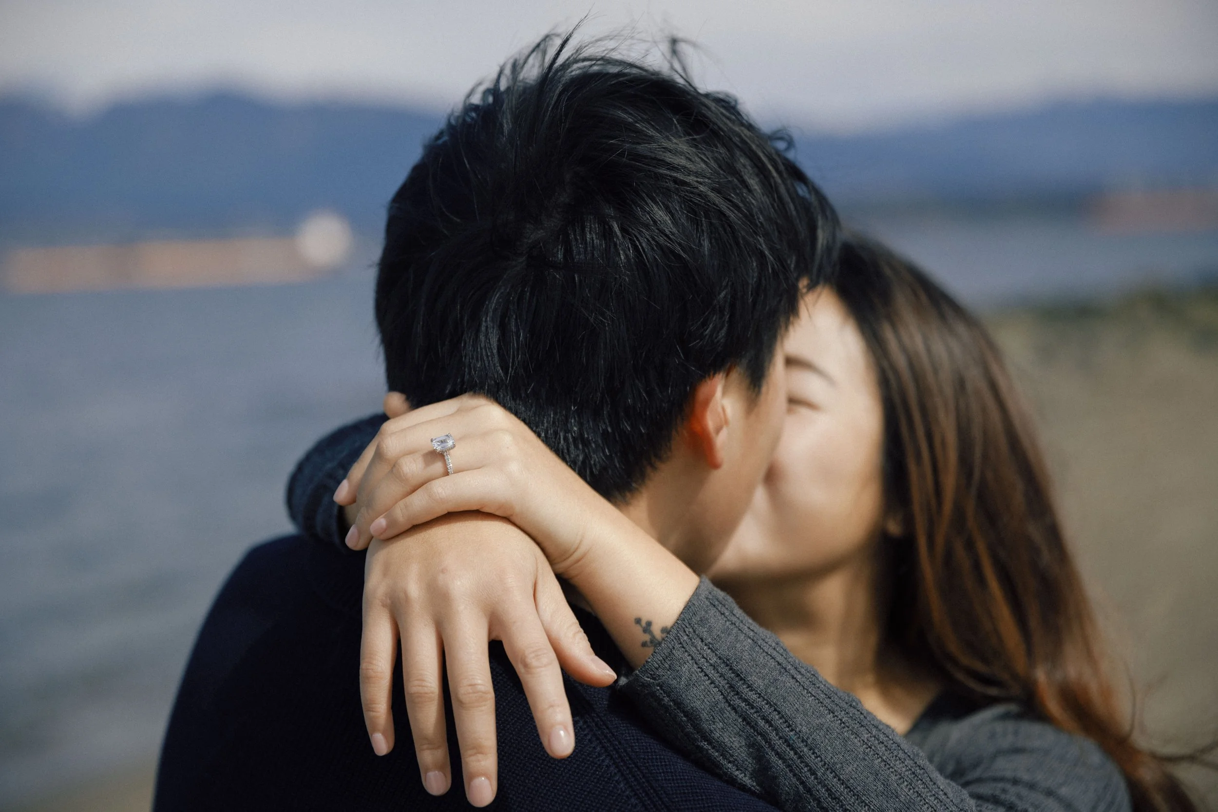 A couple embracing each other outdoors, focusing on their heads and arms, with a blurred landscape background.