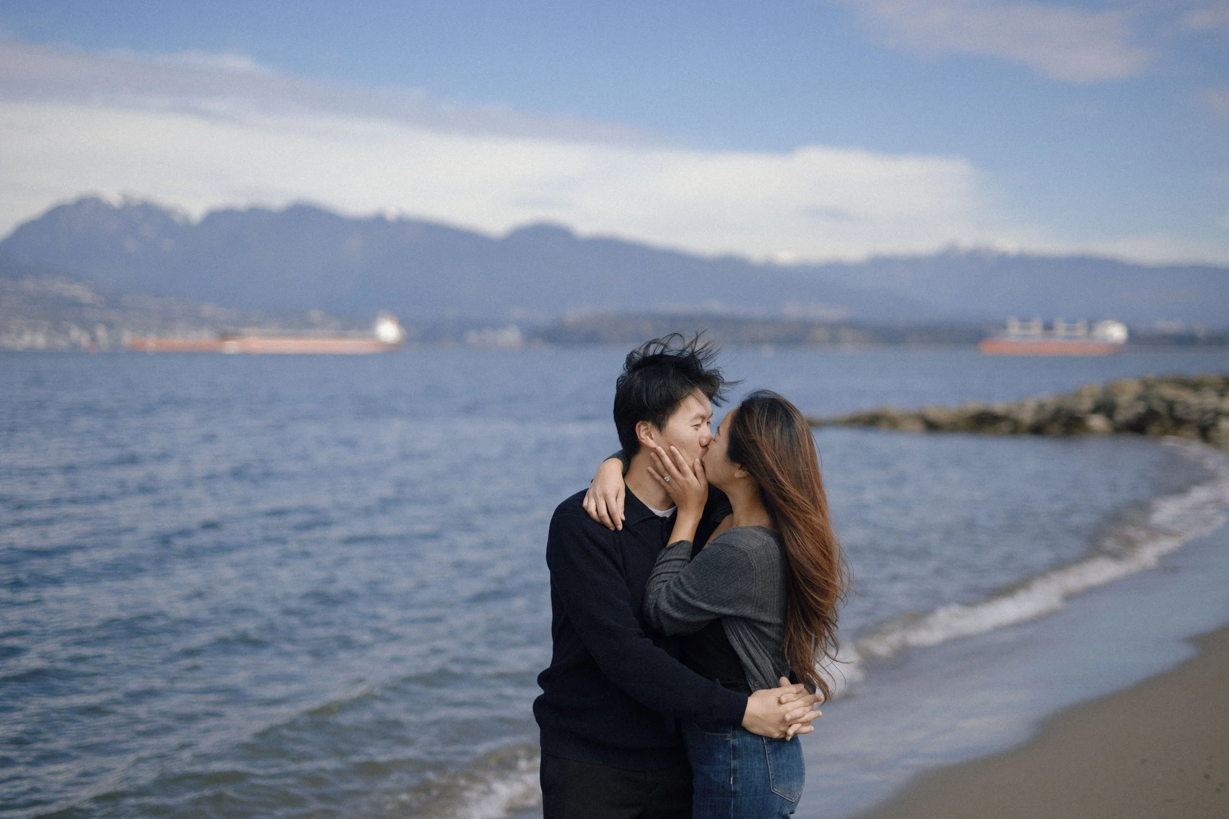 A couple is sharing a kiss on the beach, with mountains and ships in the background.