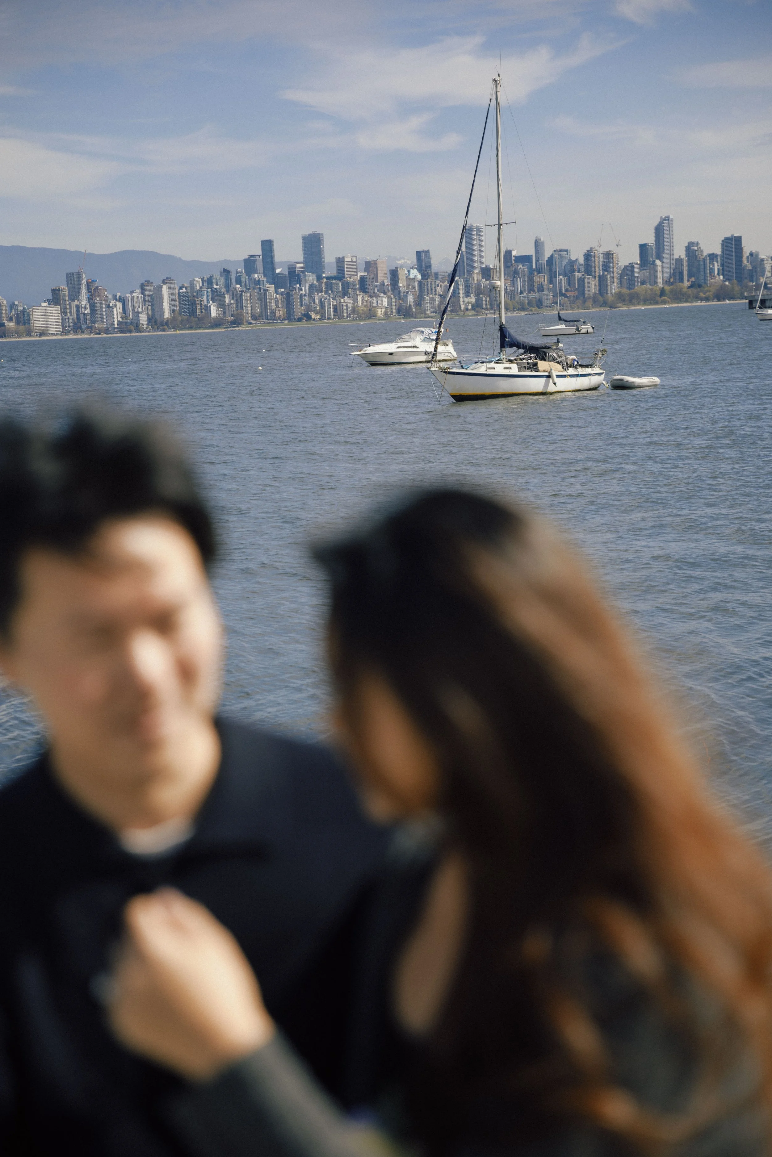 A blurred photo of people in the foreground with a city skyline and sailboats on the water in the background.