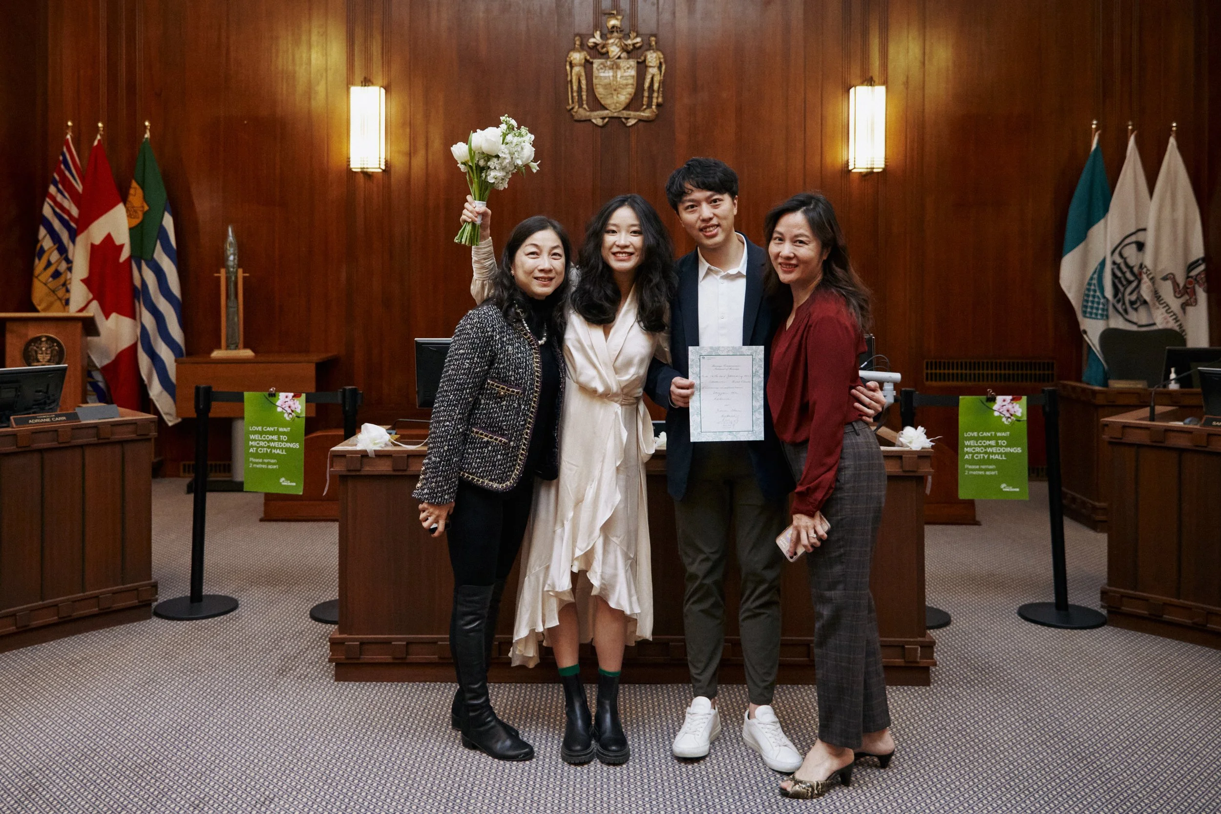 Four people stand together in a formal room, with a woman holding a bouquet of white flowers and another holding a certificate. The room features wood-paneled walls, flags, and a coat of arms. Two green signs on the desk mention a micro-wedding at ci