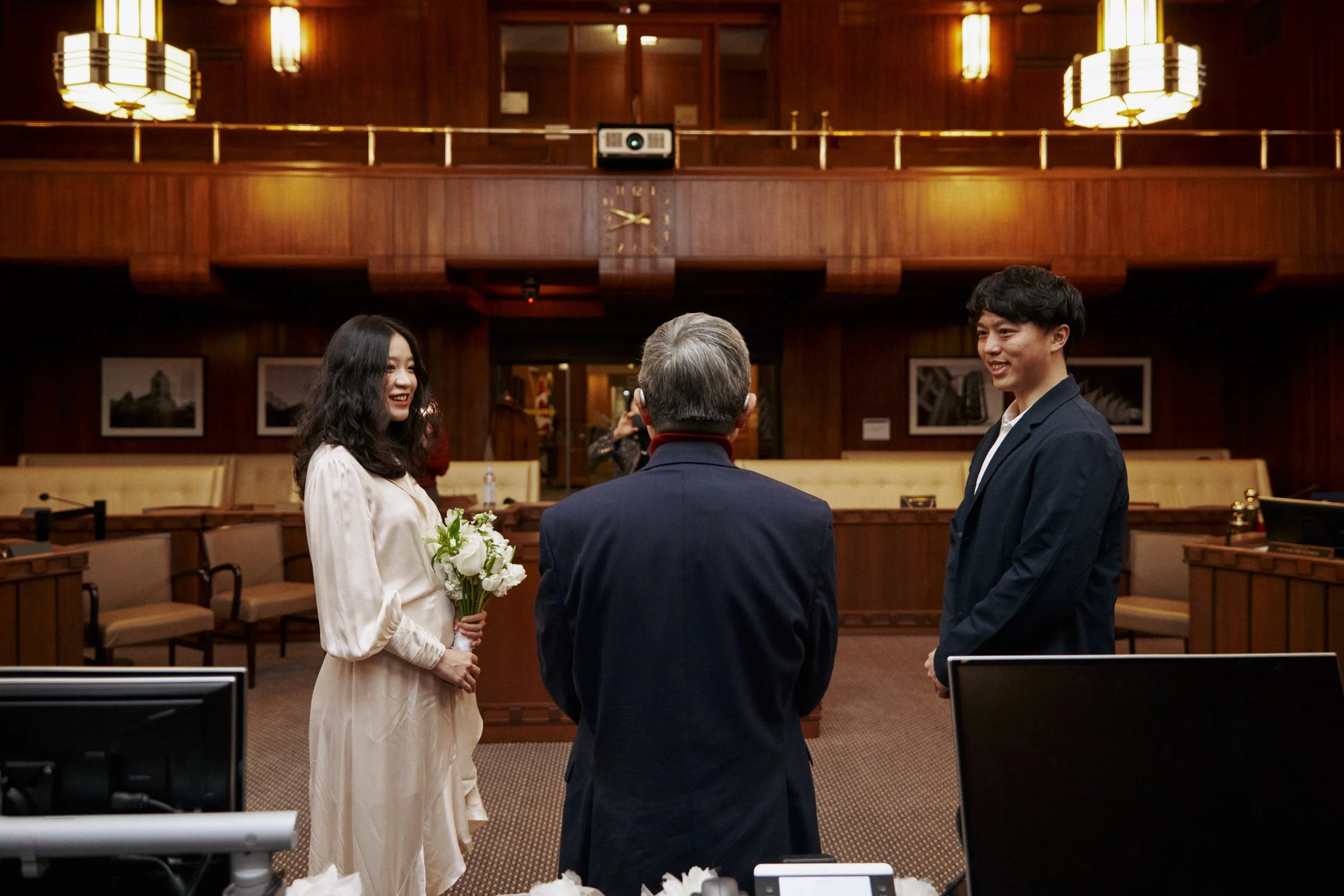 A wedding ceremony taking place in a wood-paneled room with a woman in a white dress holding a bouquet and a man in a dark suit, with an officiant standing between them. The room features warm lighting, framed pictures on the walls, and a large clock