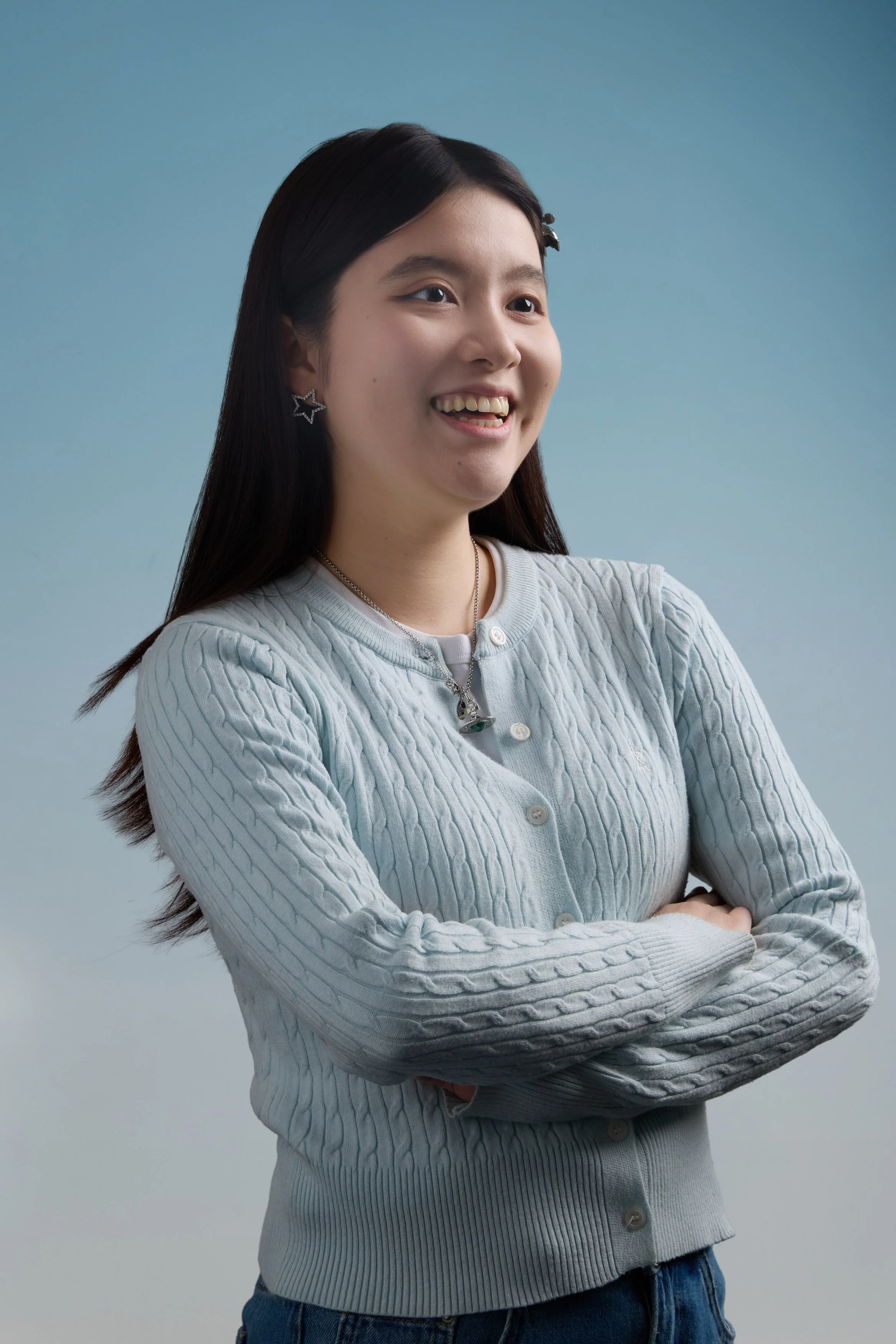 A woman with long dark hair, smiling with crossed arms, wearing a light gray cable-knit cardigan and star-shaped earrings against a light blue background.