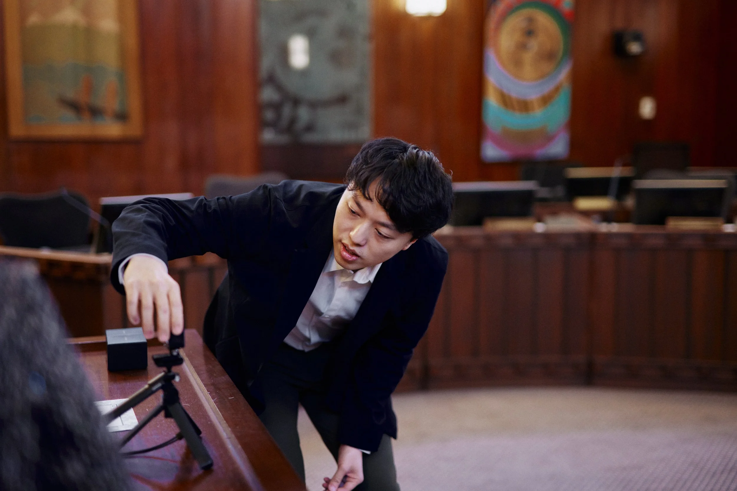 A young man with black hair, dressed in a black suit and white shirt, is leaning over a wooden table in a room with wooden paneling and colorful abstract art on the walls. He appears to be engaged in a discussion or activity with others around him.