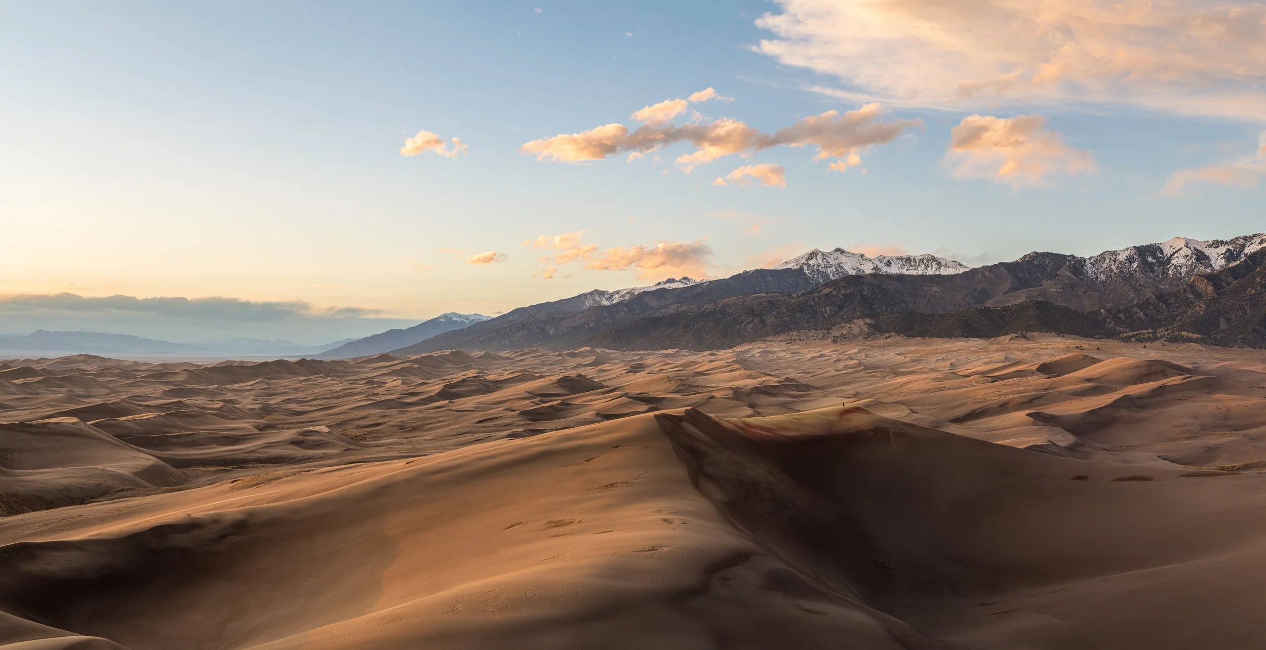 Dunes in a desert with mountains in the background and a partly cloudy sky at sunset.