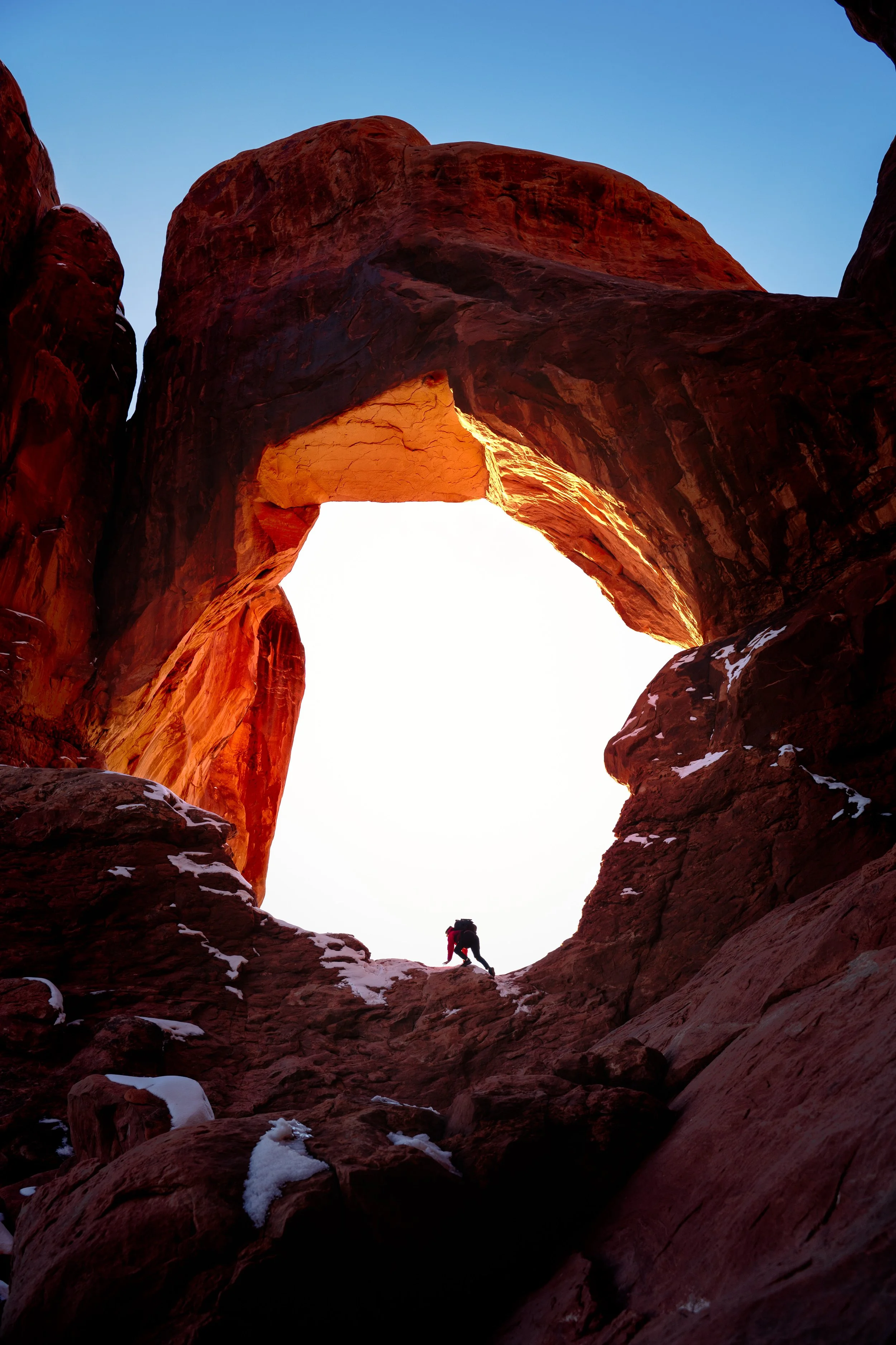 A person with climbing gear at the base of a natural rock arch formation during sunset or sunrise, with snow on the rocks.