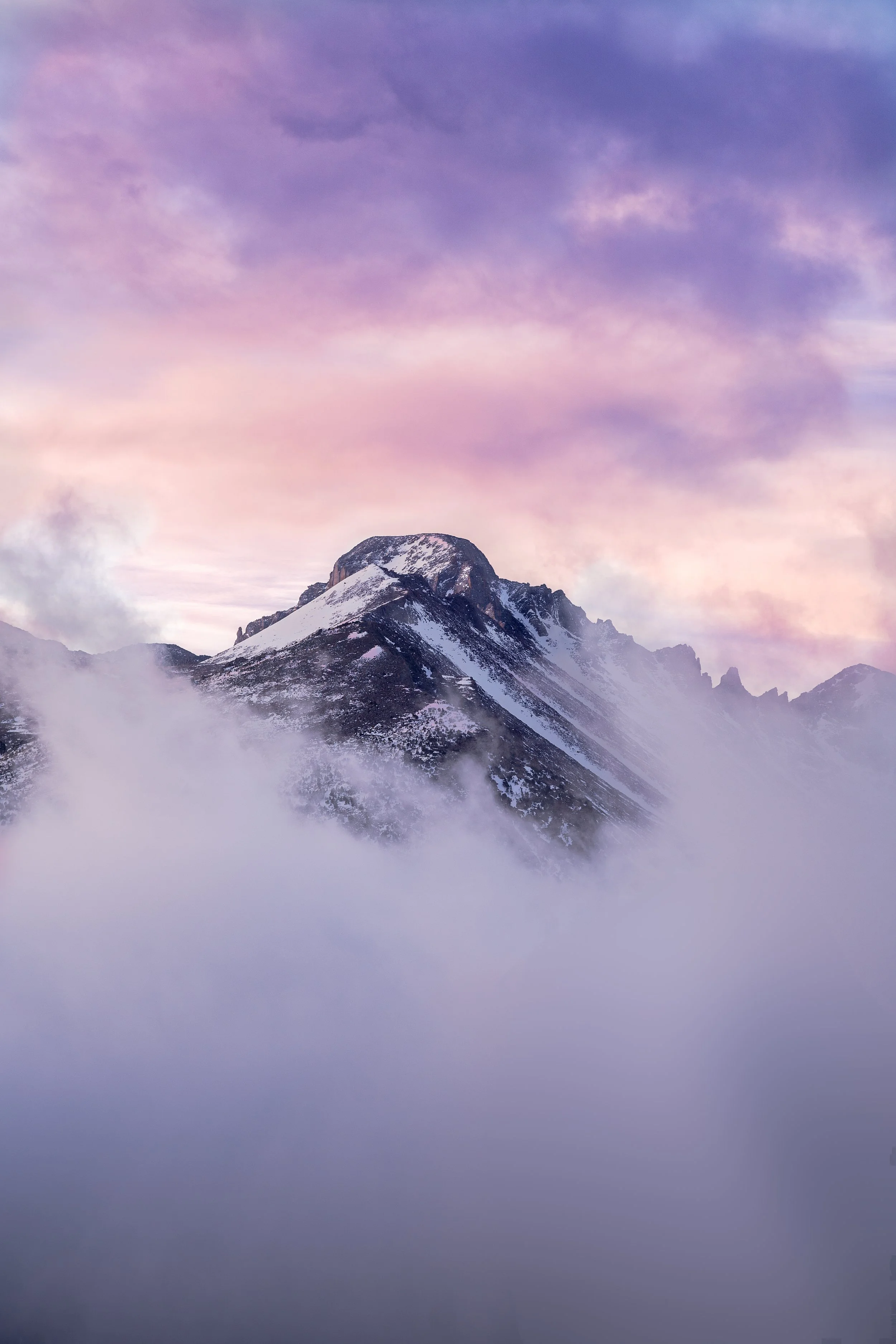 Snow-covered mountain peak surrounded by clouds during sunrise or sunset with a colorful sky.