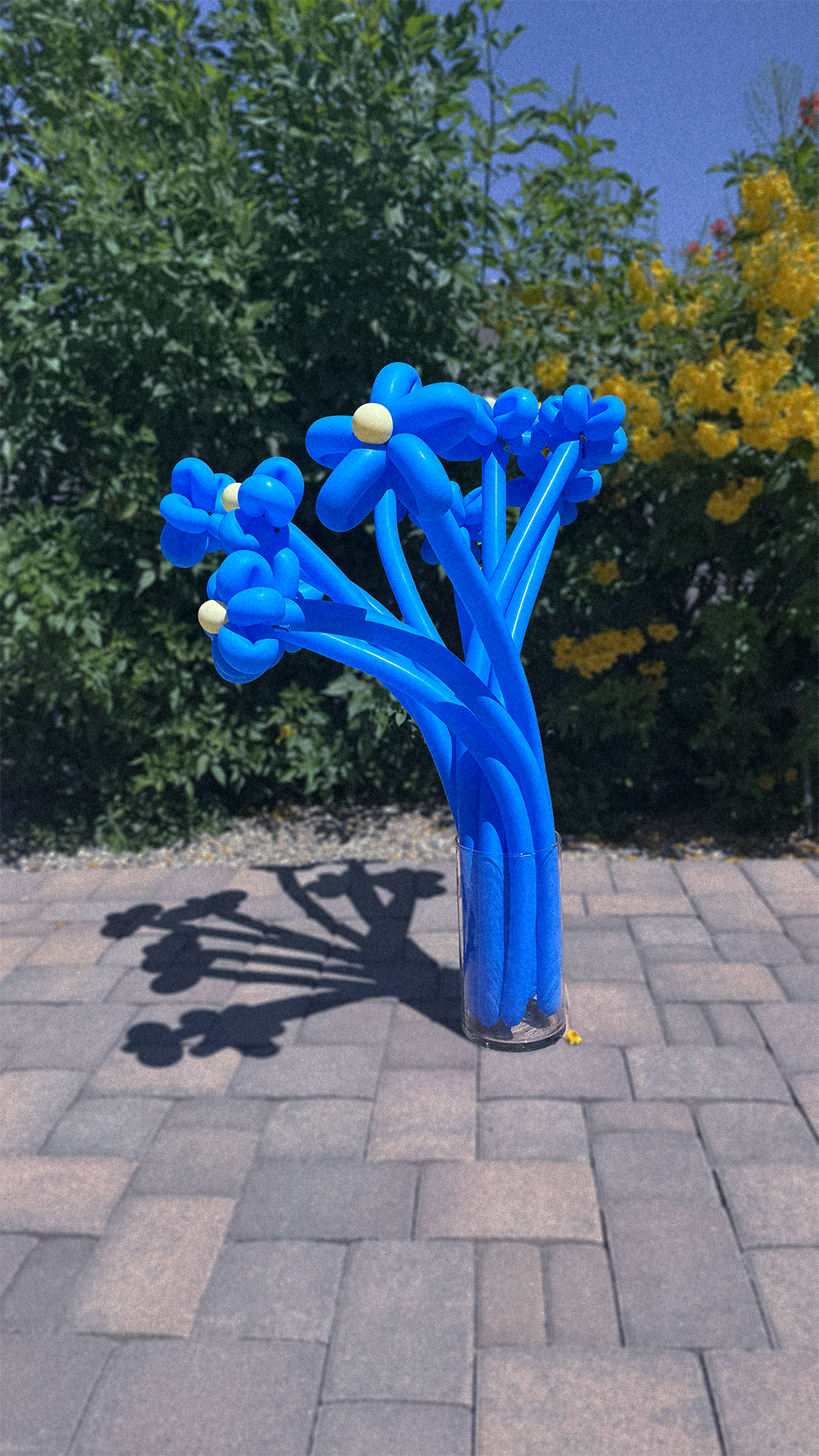 A sculpture resembling a bouquet of blue balloons in the shape of flowers, placed in a clear glass vase, casting a shadow on the brick pavement in an outdoor garden setting at dusk.