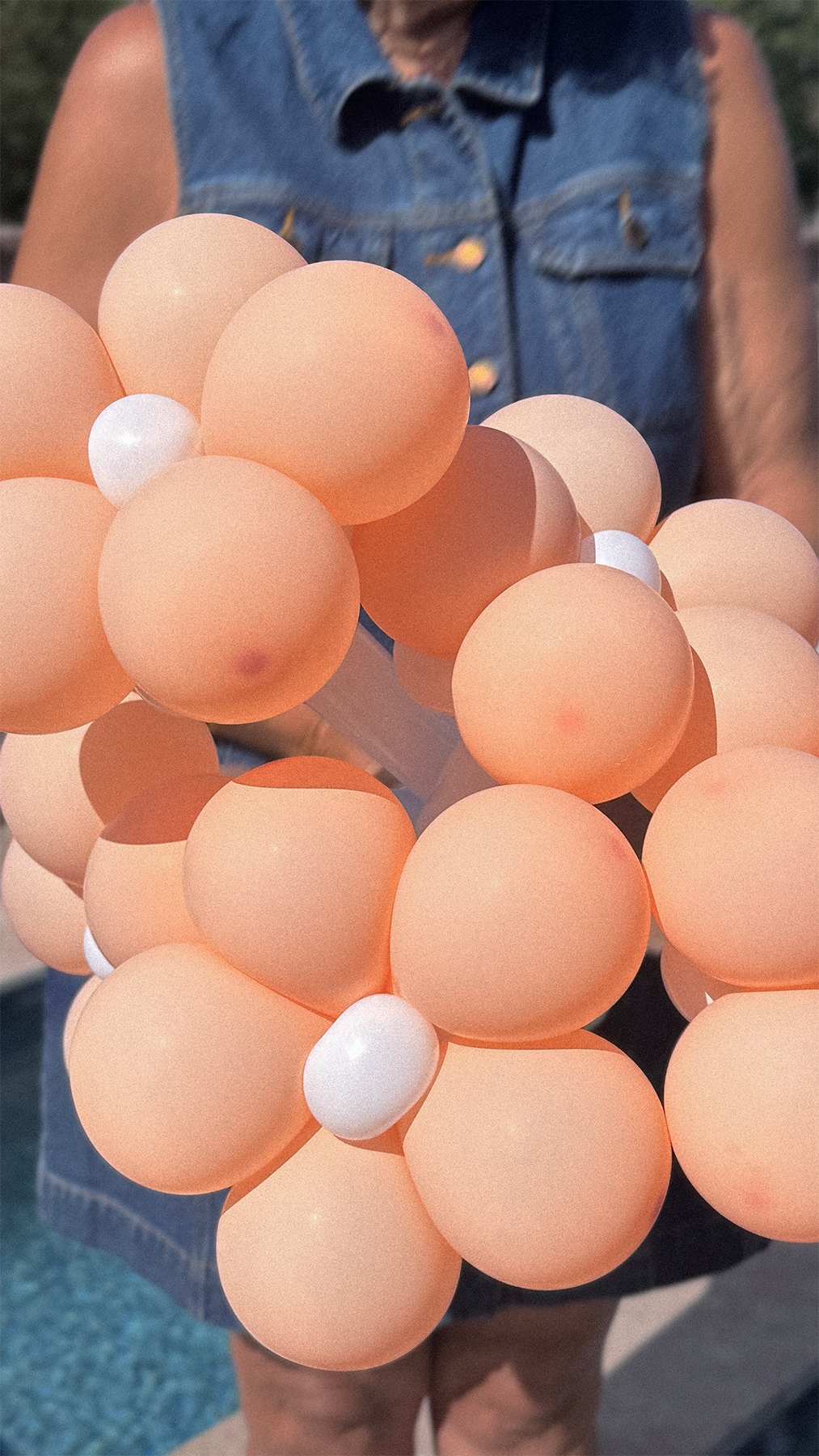 Person holding a bunch of peach-colored balloons with a few white balloons, wearing a denim vest.