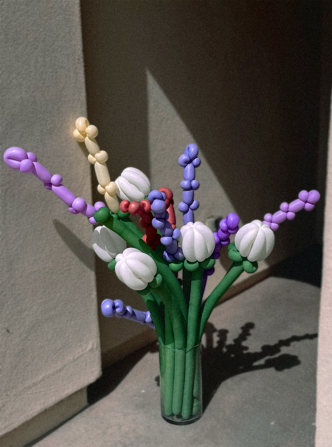 A colorful artificial flower bouquet with purple, white, red, and blue flowers in a clear glass vase, placed on a gray surface near a wall.