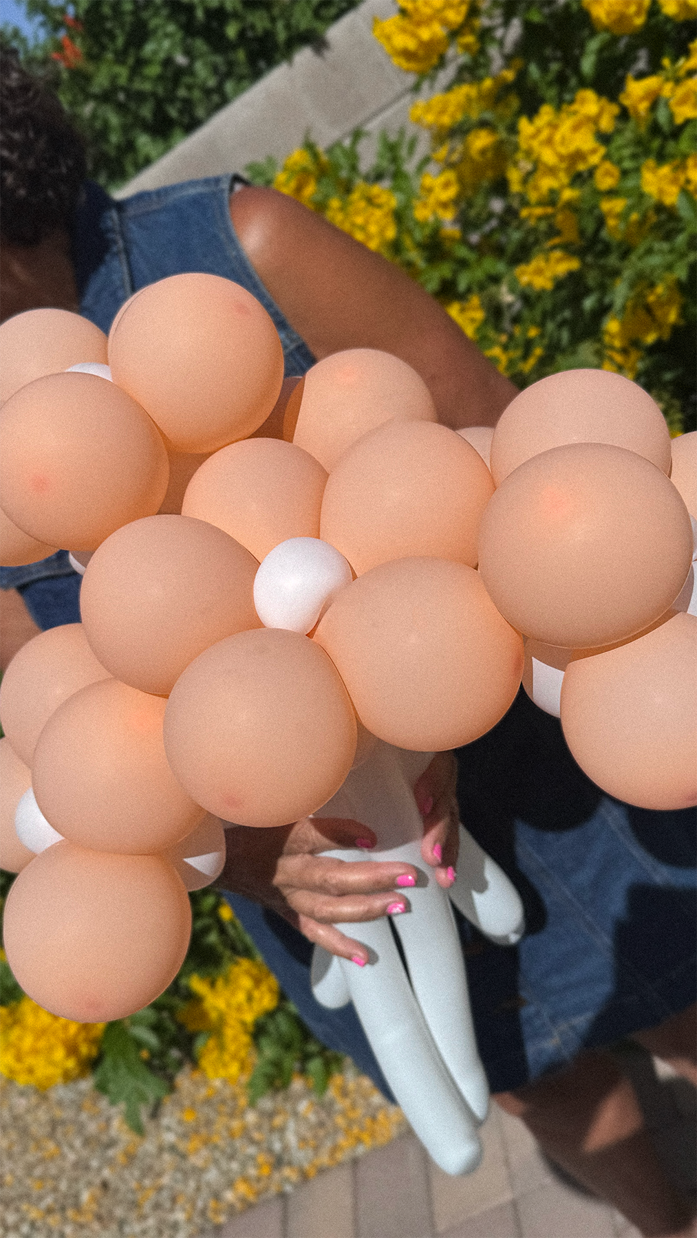 Person holding a bunch of beige, white, and black balloons outside near yellow flowers and greenery, with a person wearing a denim vest and pink nail polish.