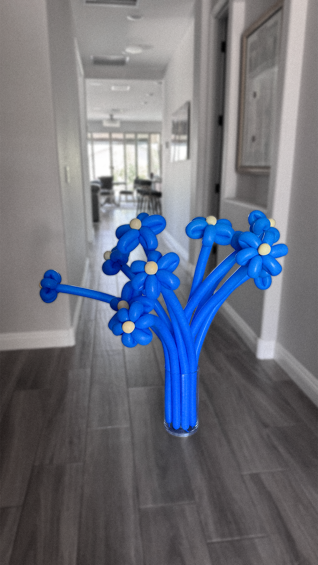 A blue balloon bouquet resembling flowers in a clear vase, placed on a wooden floor in a hallway.