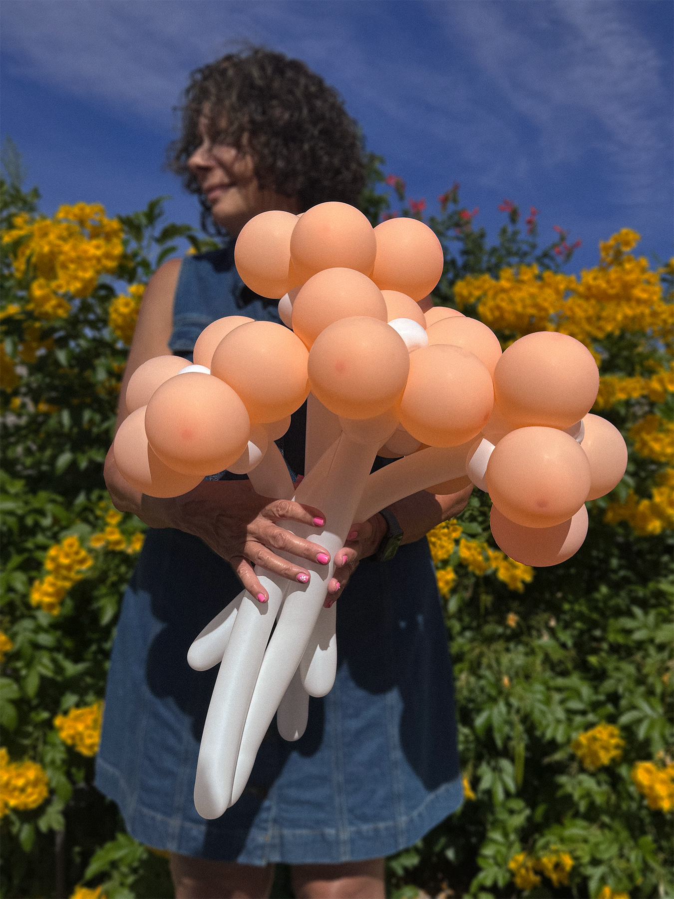 A woman with curly hair holding a bouquet of balloon-like objects shaped like eggs in an outdoor garden with yellow flowers and a blue sky.