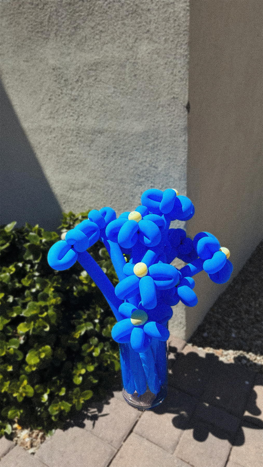 Blue balloon sculpture resembling a bouquet of flowers placed outdoors on a sunny day, with greenery and concrete surfaces in the background.