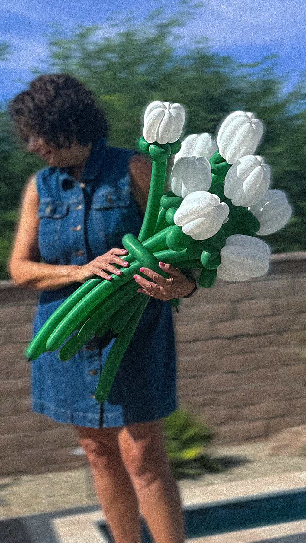 A woman holding a bunch of balloon tulips with white petals and green stems in an outdoor setting, wearing a sleeveless denim dress and standing on a wooden deck.