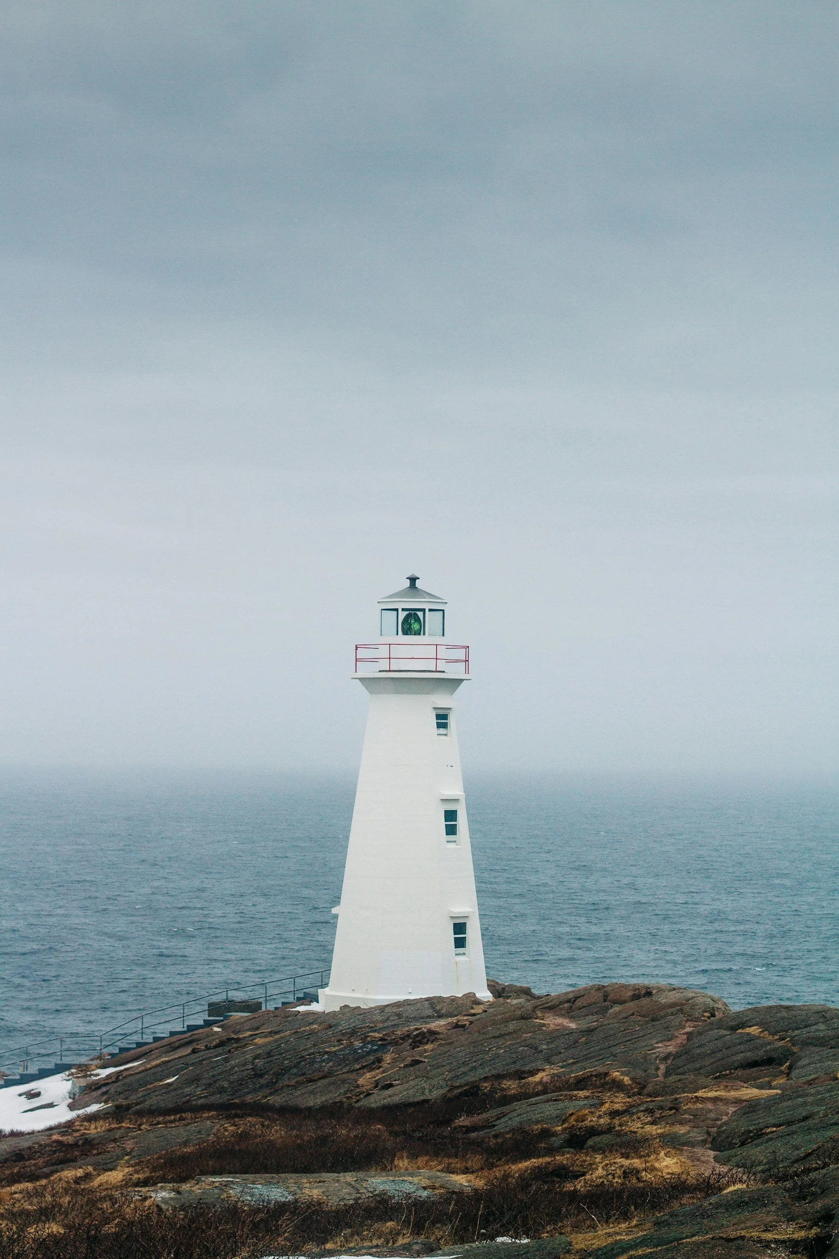 White lighthouse on rocky shoreline with overcast sky and ocean in the background.
