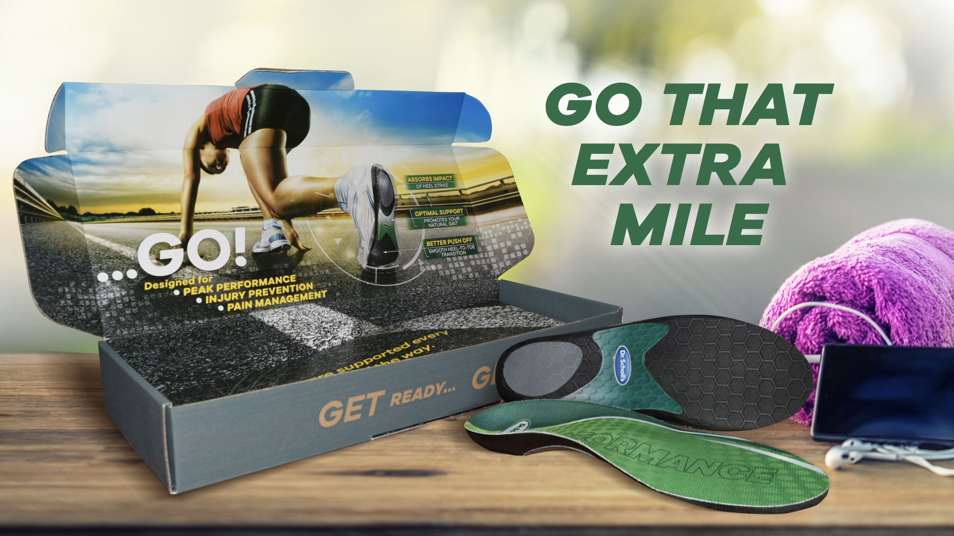 A sports shoe insoles display with the packaging nearby and a pink towel, a smartphone, and earphones on a wooden surface. The background is blurred with a slogan in green bold text that reads 'GO THAT EXTRA MILE'.