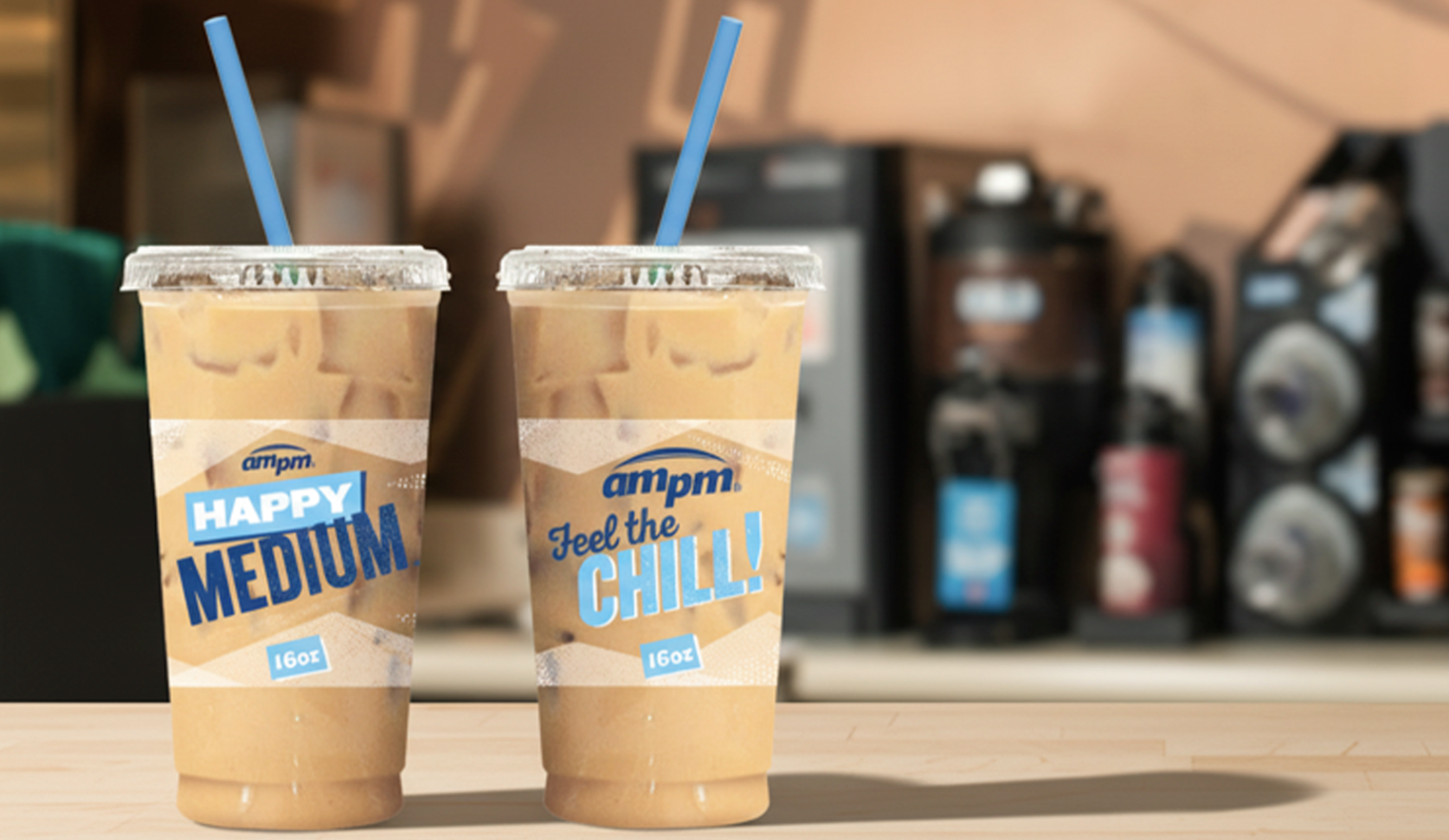 Two iced coffee drinks in plastic cups with blue straws, labeled 'Happy Medium' and 'Feel the Chill,' placed on a light wood countertop in a coffee shop setting.