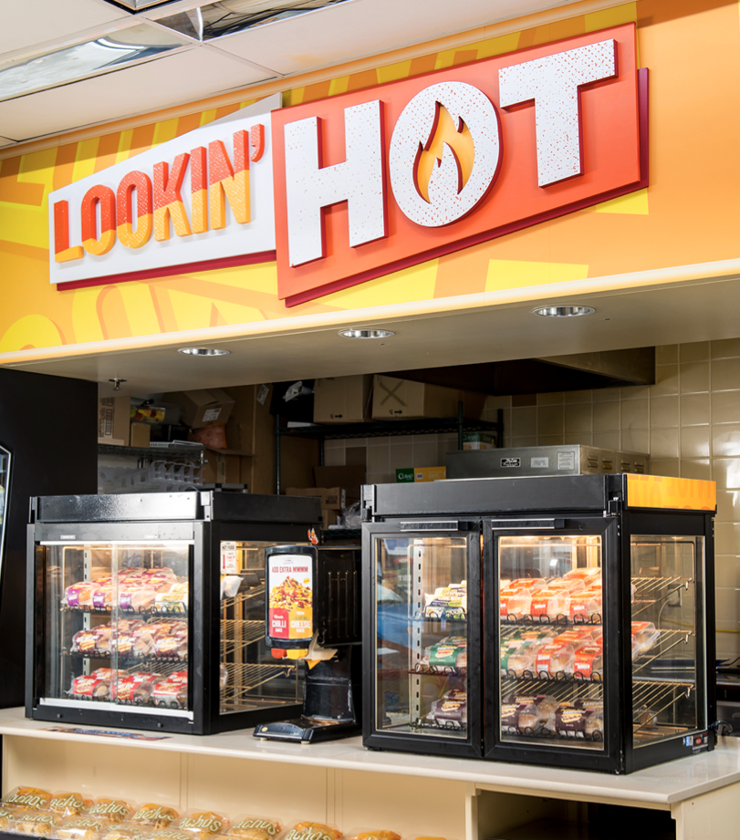Store signage reading "Lookin' Hot" with a stylized flame inside the letter "O". Two glass display cases filled with packaged hot food items are underneath the sign.