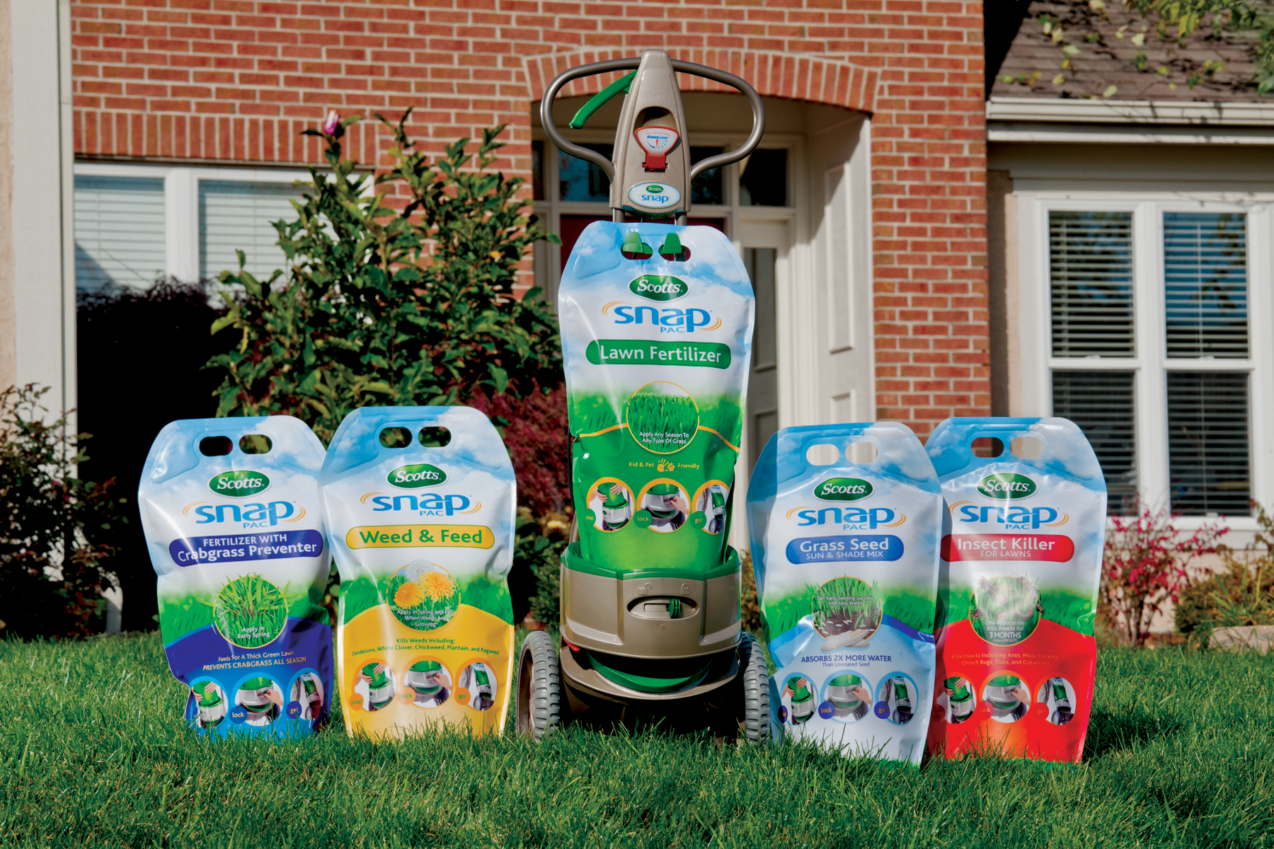 Lawn care sprayer surrounded by five bags of Scotts Snap lawn fertilizer products on a front lawn with a brick house in the background.