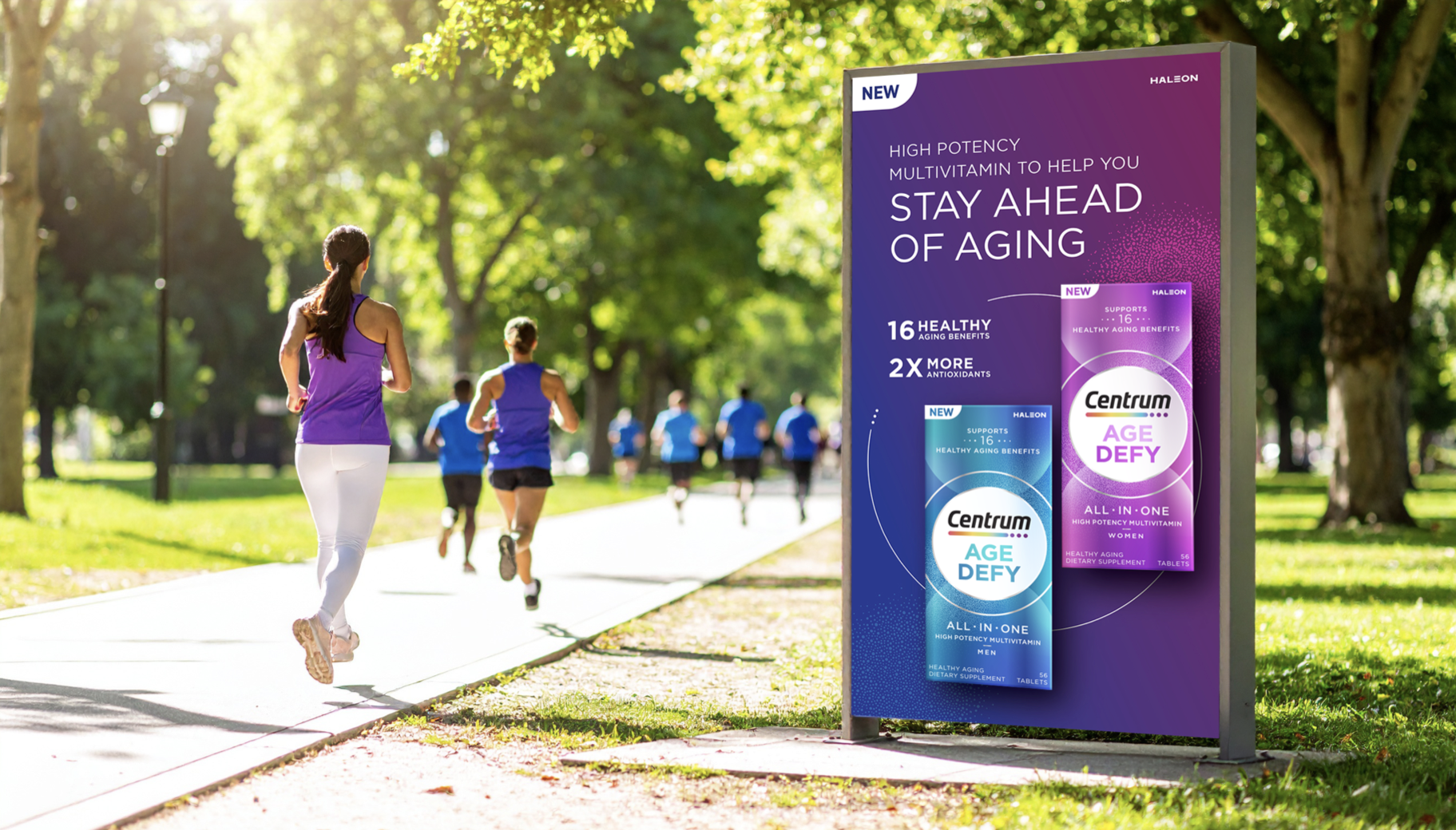 An outdoor scene in a park with sunlight filtering through green trees, a woman jogging in a purple tank top and white leggings, and other people running on the paved path. There is a purple advertisement board for Centrum multivitamins for aging health benefits.
