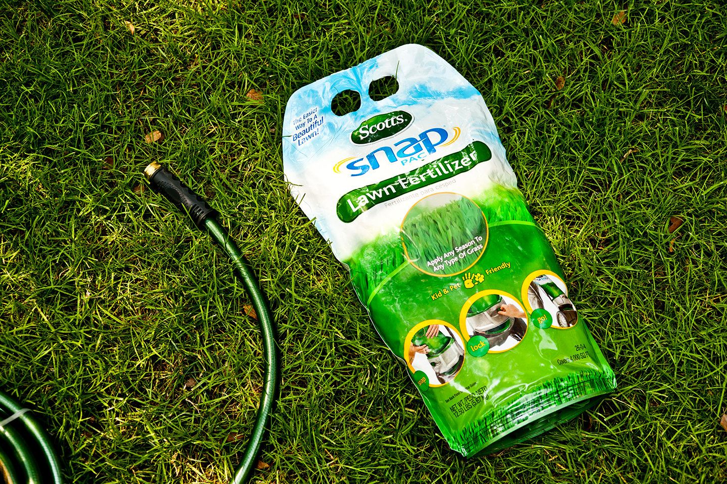 A bag of Scotts Snap Pac lawn fertilizer lying on green grass with a garden hose nearby.