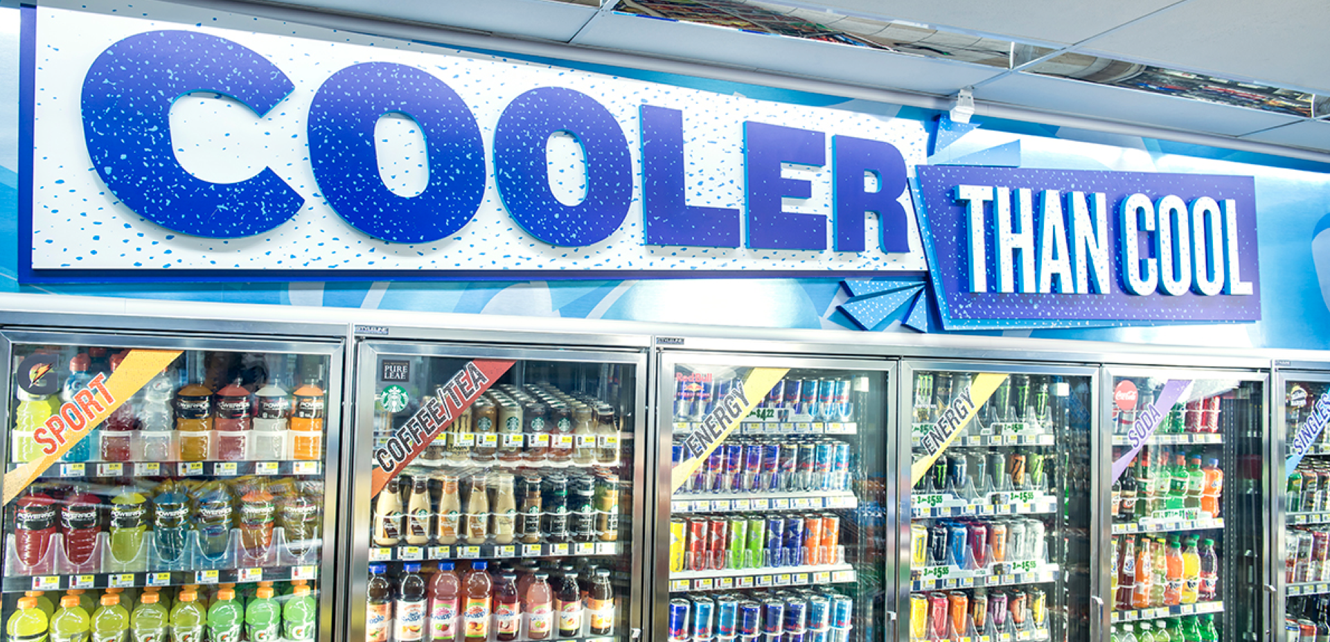 A store freezer with a blue sign that says "Cooler Than Cool". Inside the freezer, various beverages are visible, including sports drinks, coffee and tea, energy drinks, soda, and single-serve bottles.