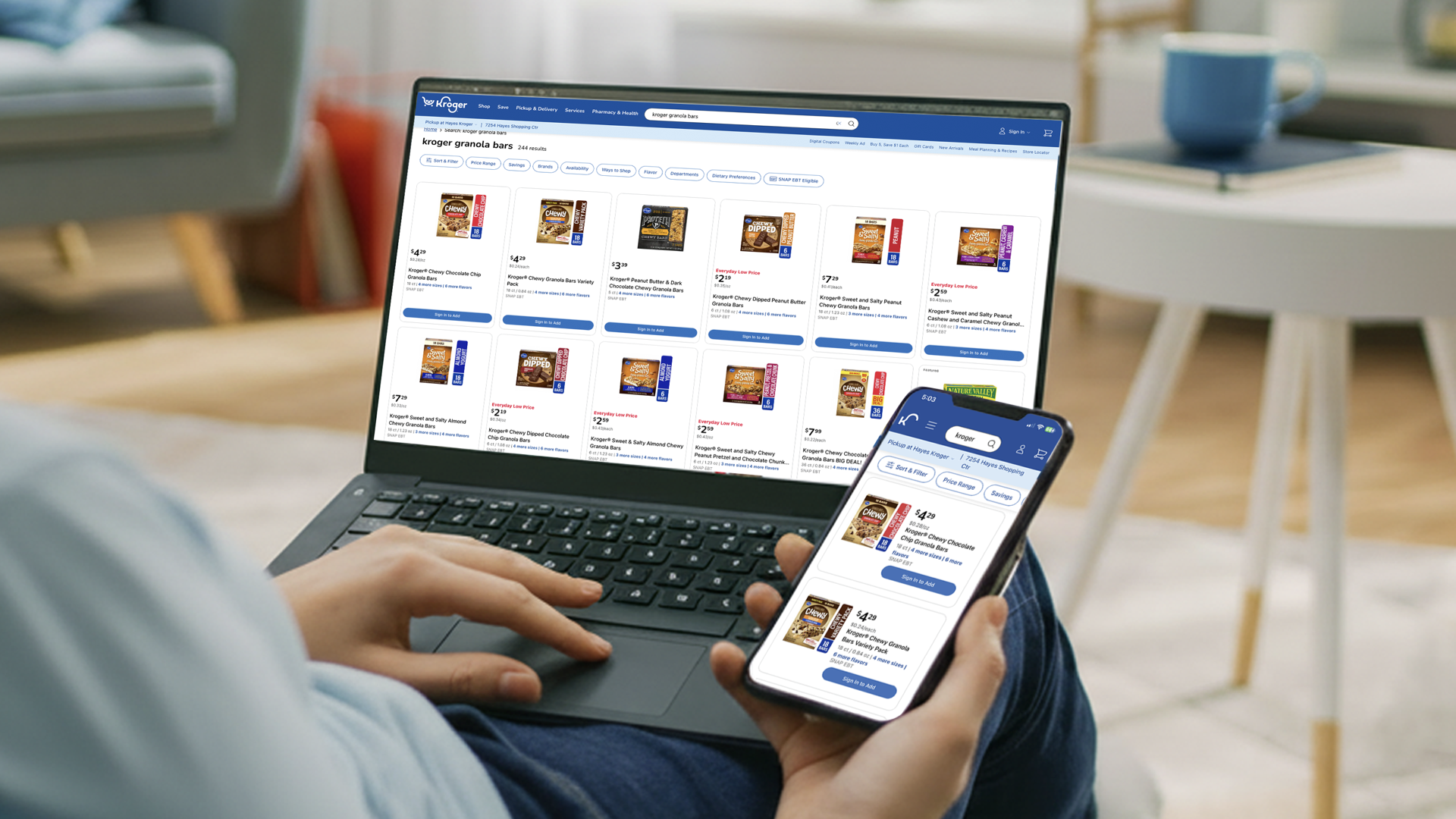 Person sitting on a chair using a laptop and smartphone to shop for grocery items online, displaying various granola bars on the screens.