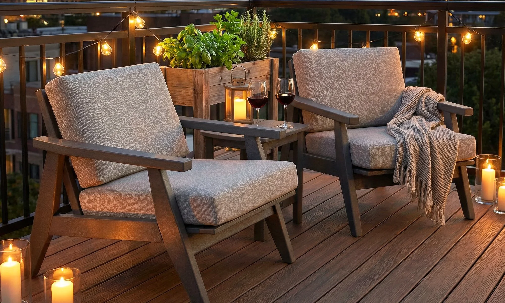 A cozy outdoor balcony patio at dusk with two cushioned chairs, a small table with two glasses of red wine, a lantern, candle candles, string lights, and potted plants.