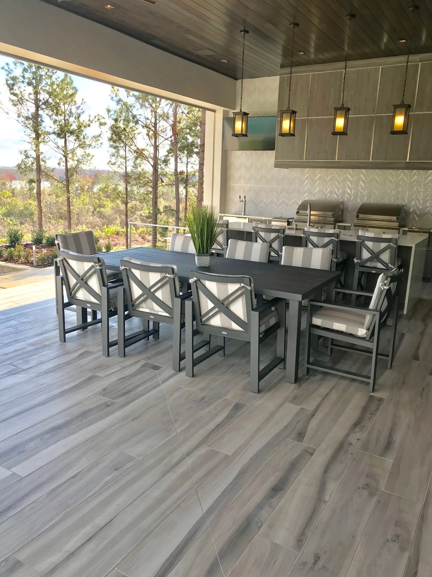 Indoor dining area with a dark wood table, white and gray chairs, a potted plant centerpiece, large window showing trees outside, and a kitchen with metallic grills and hanging pendant lights.