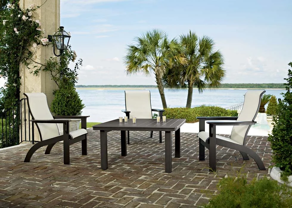 Outdoor patio with three white and black chairs around a black table, overlooking a water body with two palm trees and a partly cloudy sky.