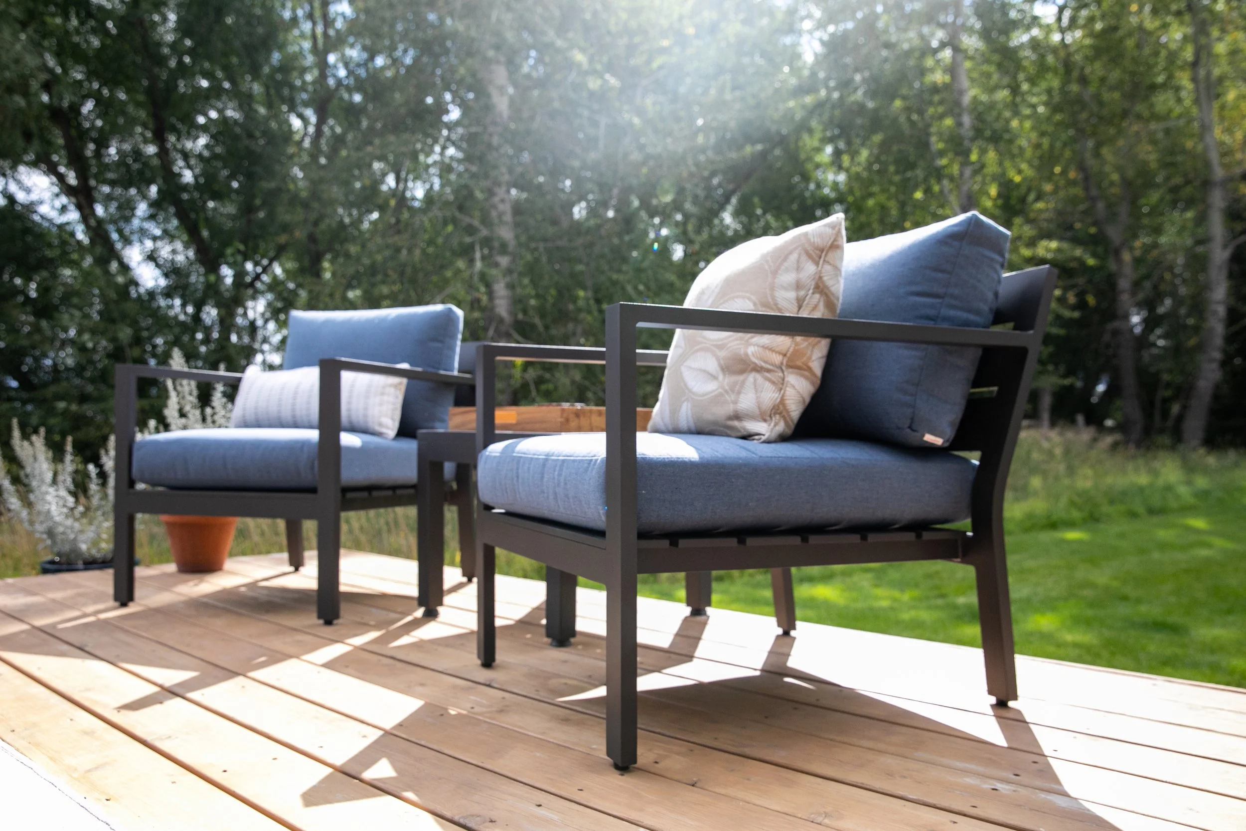 Outdoor patio with beige cushioned seating including a sofa and two armchairs, a black coffee table with a potted plant, two lanterns, a large area rug, and a scenic background of trees and houses.