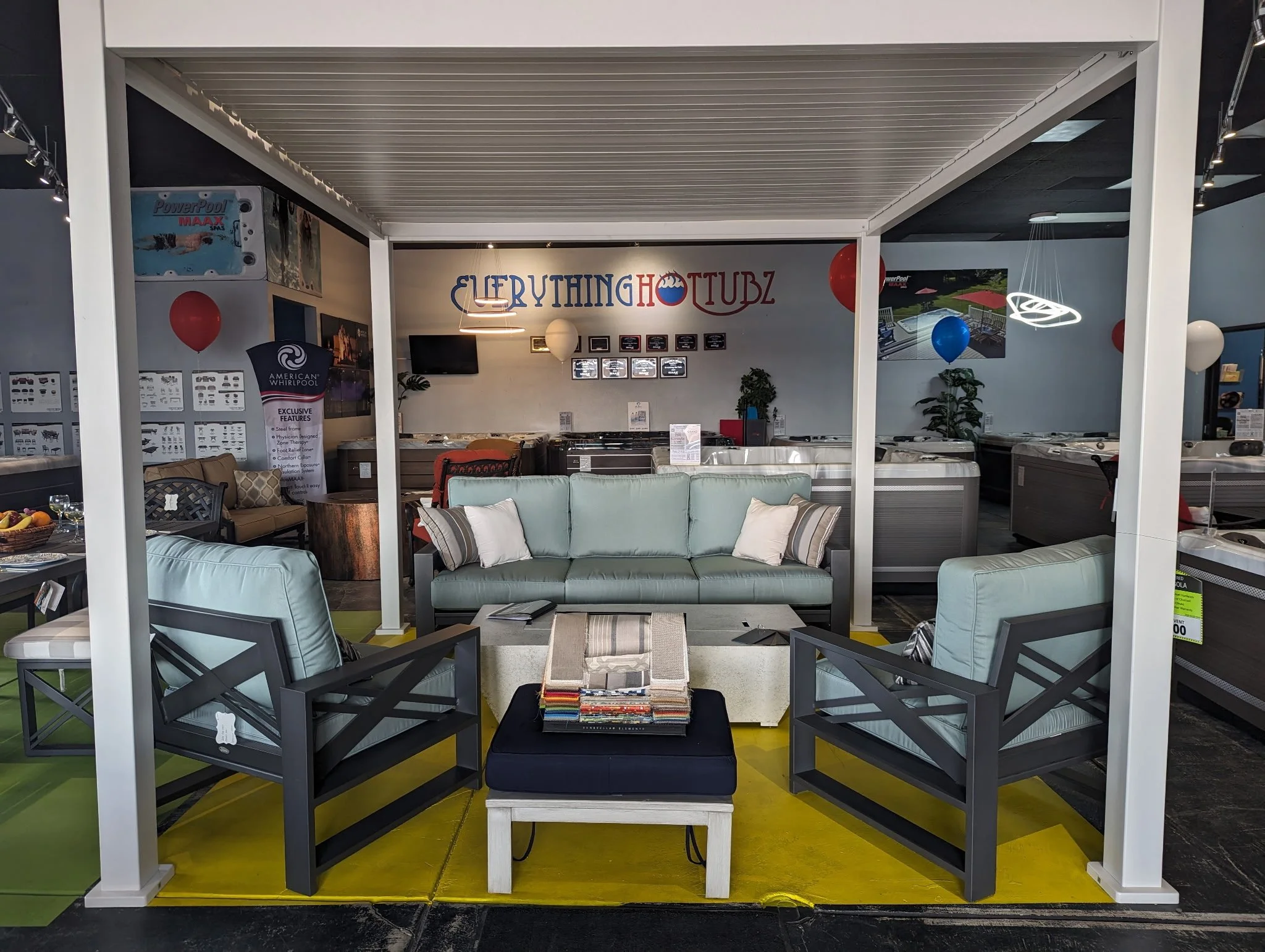Indoor furniture display with a light green sofa, two matching armchairs, a black ottoman with fabric samples, and balloons, inside a store named Everything Hot Tubz.