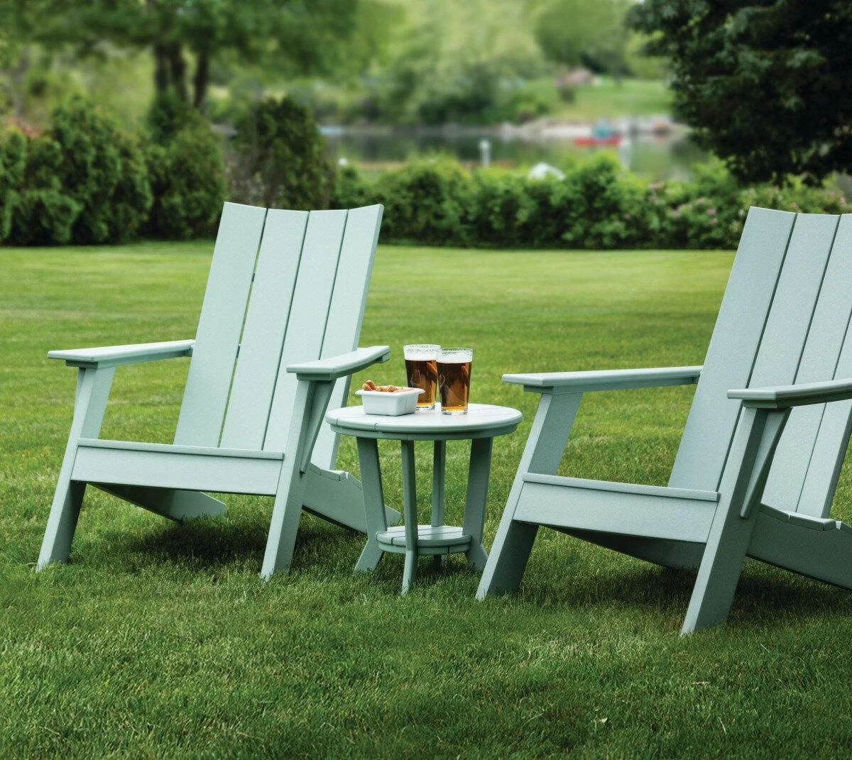 Two Adirondack chairs with a small round table between them outdoors, with three glasses of beer and a bowl of snacks on the table, on a grassy lawn with trees and a lake in the background.