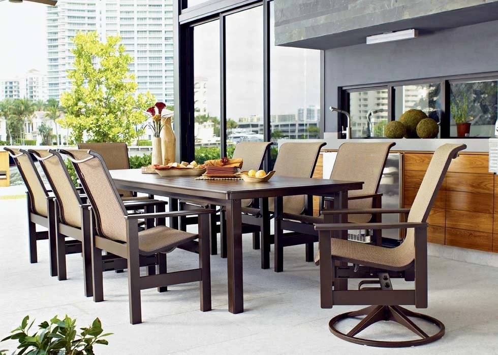 Modern outdoor patio dining area with a large rectangular wooden table, surrounded by ten beige cushioned chairs, on a balcony with cityscape view, glass windows, and a sideboard with decorative items.