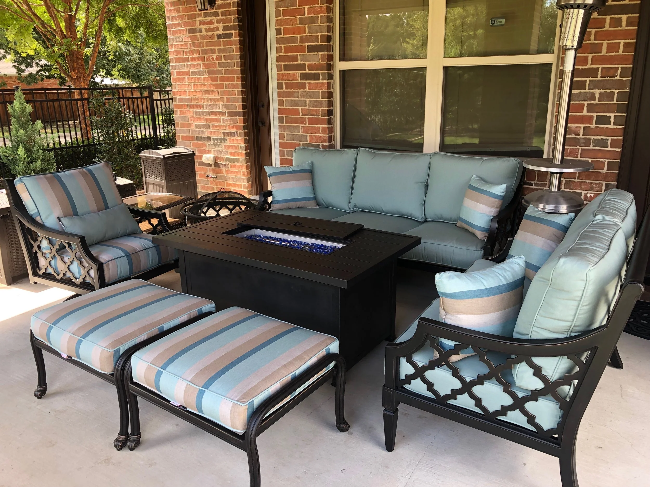 Outdoor patio furniture set with light blue cushions, striped pillows, a black fire pit table, and a patio heater in front of a brick house wall with a large window. There are trees and greenery in the background.