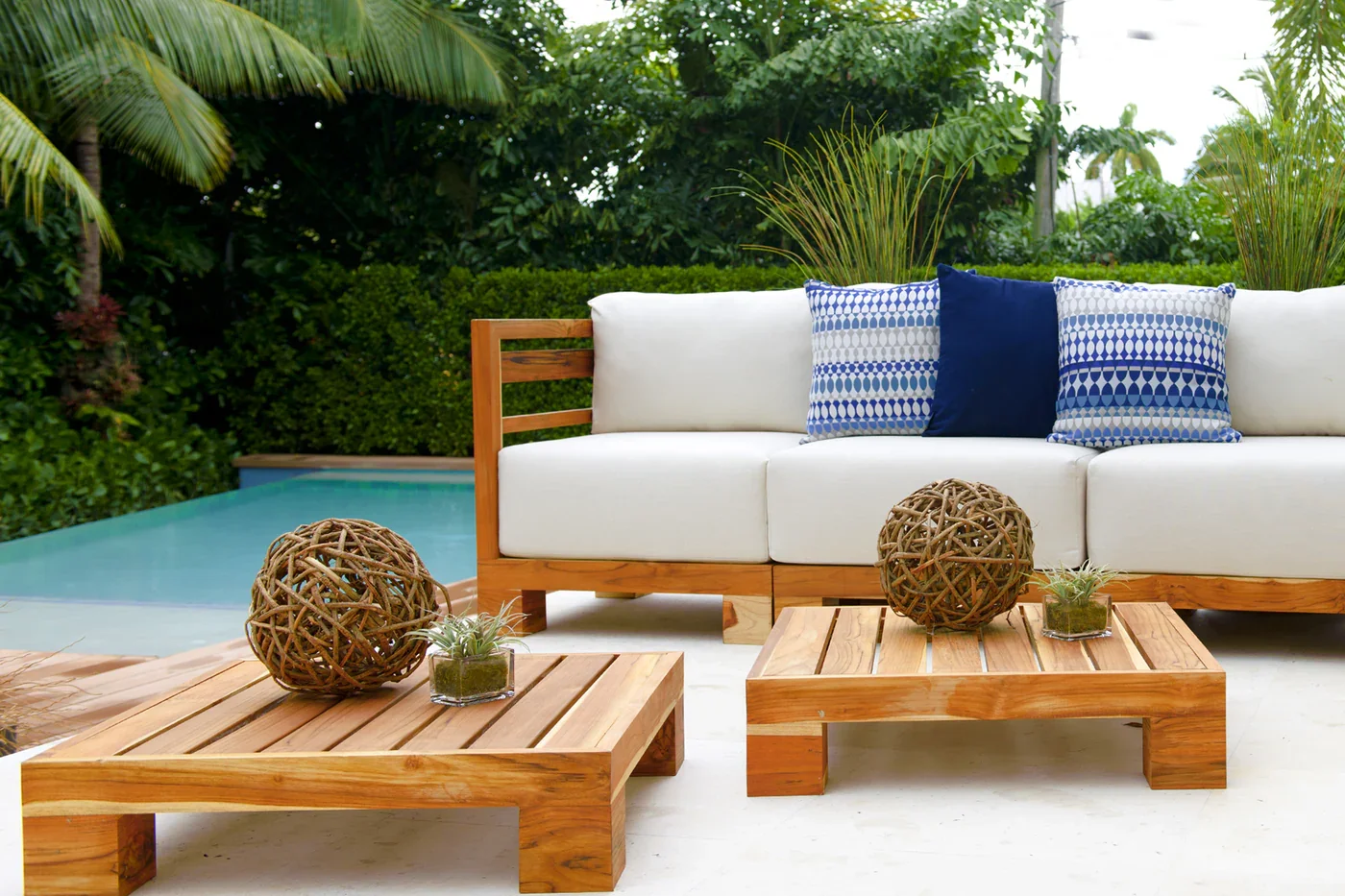 Outdoor patio with white sofa, blue patterned pillows, and wooden coffee tables surrounded by greenery and a swimming pool in the background.