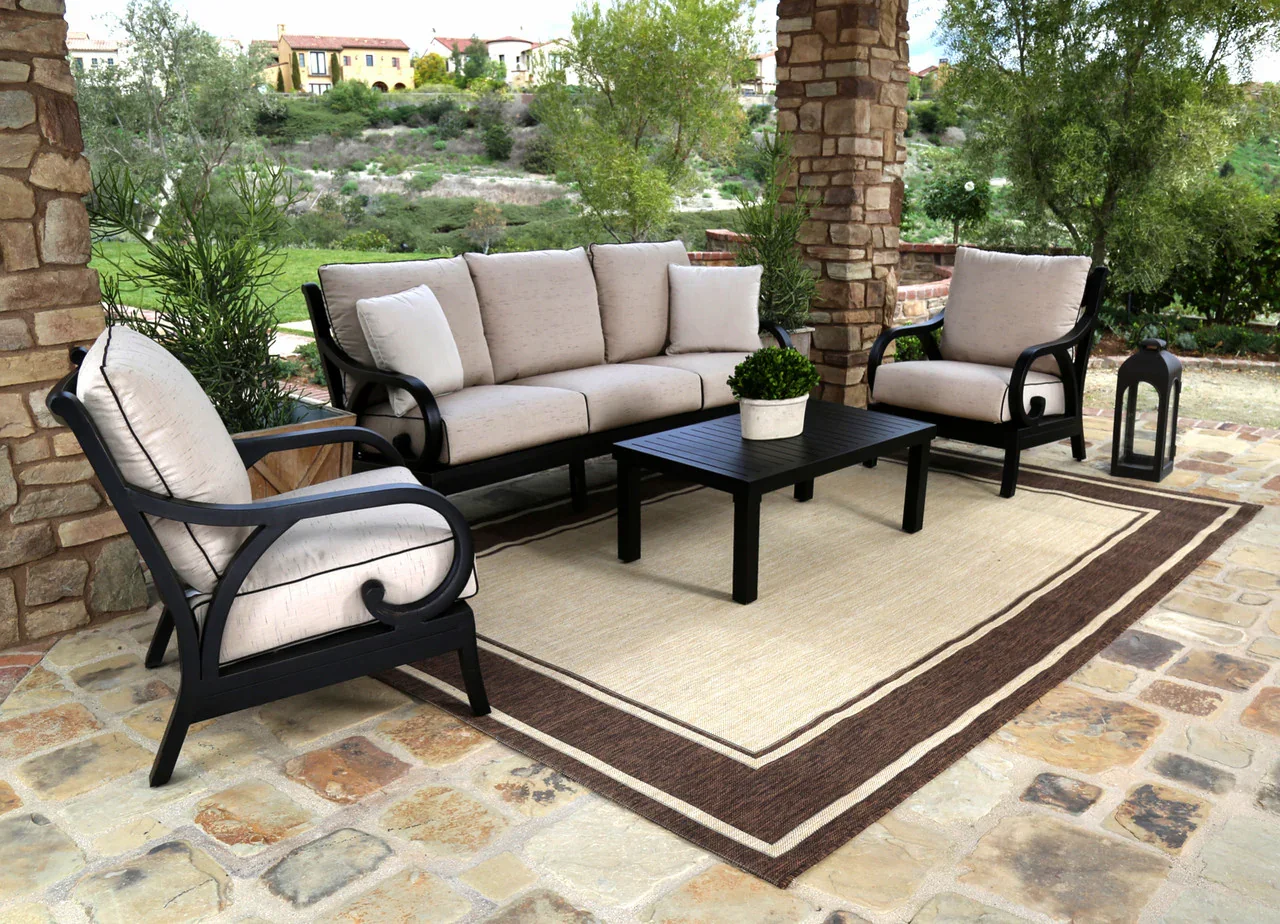 Outdoor patio with beige cushioned seating including a sofa and two armchairs, a black coffee table with a potted plant, two lanterns, a large area rug, and a scenic background of trees and houses.