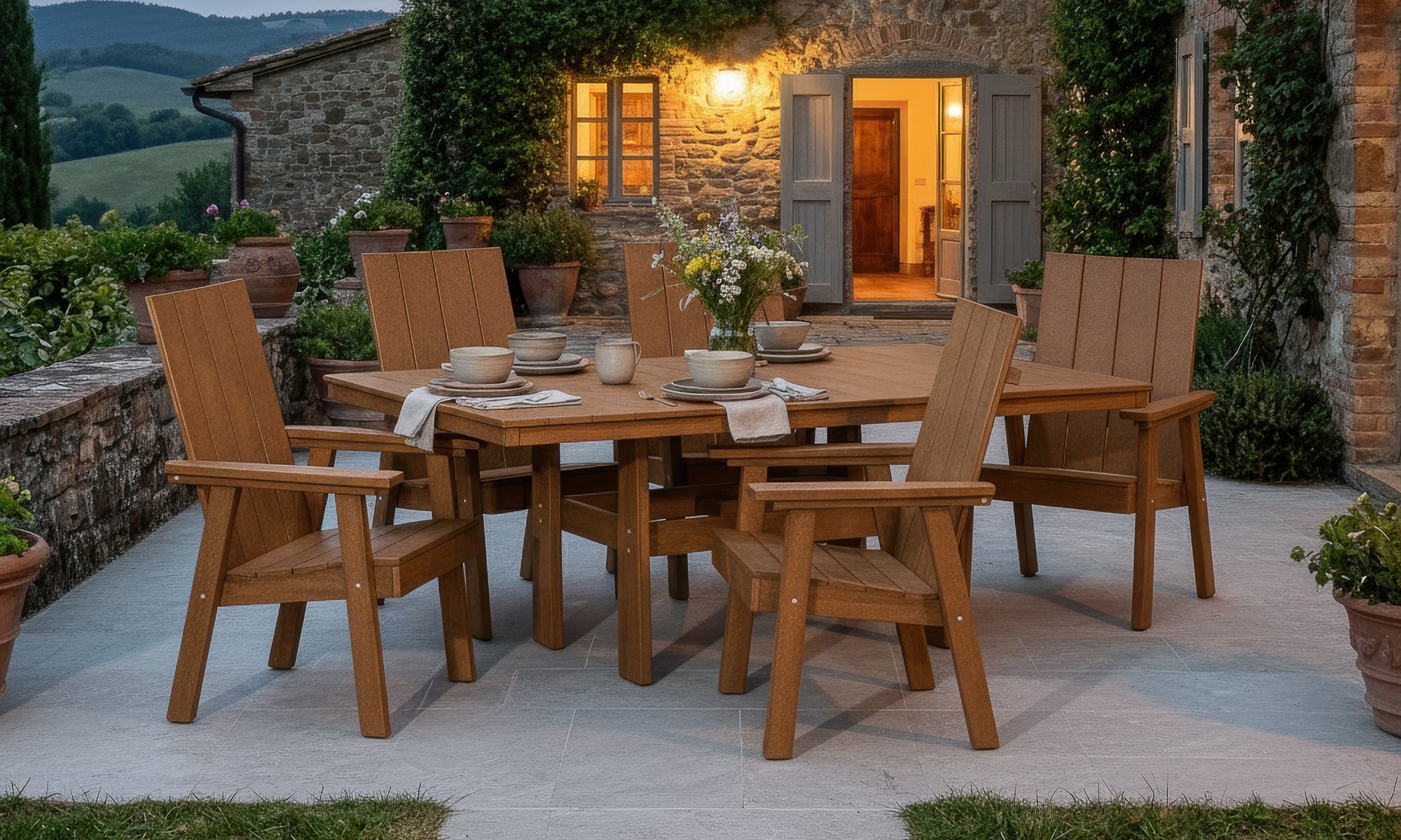 An outdoor patio with a wooden dining table set for six, surrounded by six matching wooden chairs with armrests. The table is decorated with a vase of flowers and tableware including bowls, plates, and napkins. In the background, a stone house with open door and windows, warm interior lighting, and lush greenery, including potted plants and climbing vines, is visible. The setting appears to be during the evening.