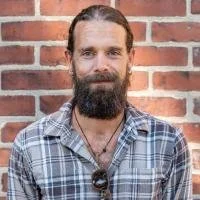 Portrait of a man with a beard wearing a plaid shirt, standing in front of a brick wall.