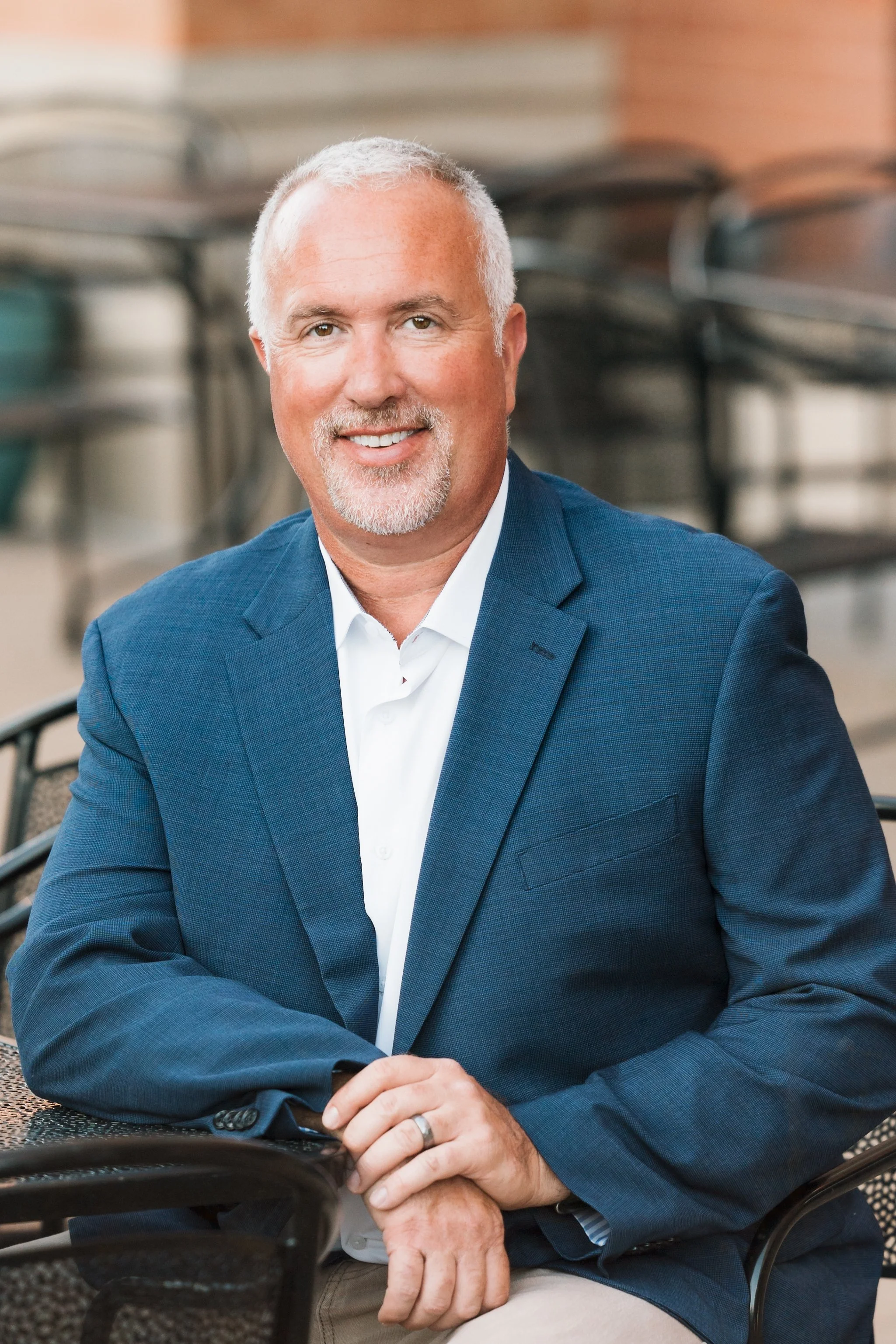 A middle-aged man with short gray hair, a beard, and a friendly smile, sitting outdoors at a table in a public area, wearing a blue blazer over a white shirt.