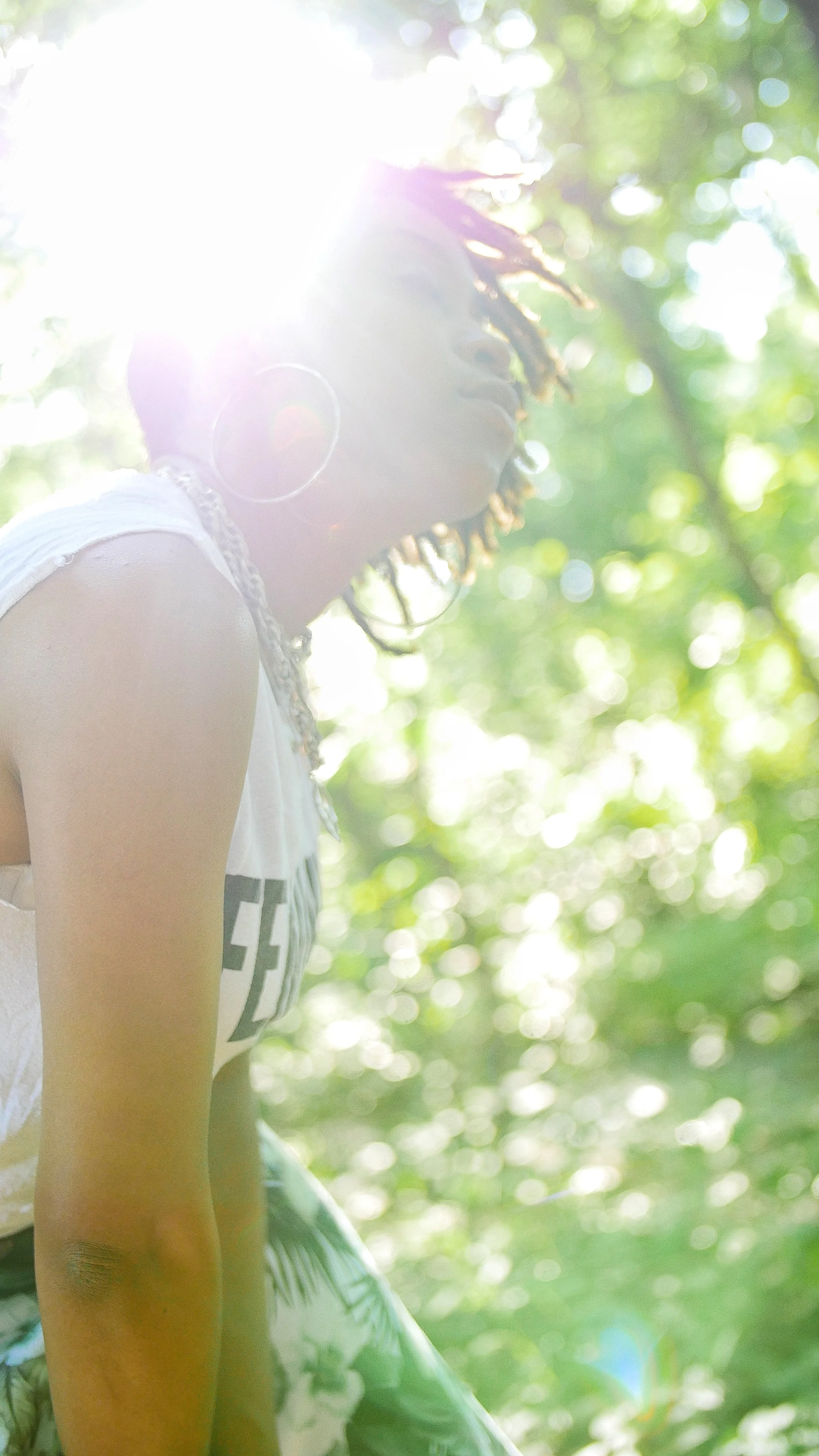 Young woman with dreadlocks wearing large hoop earrings, sitting outdoors in sunlight filtering through trees, with bokeh background.