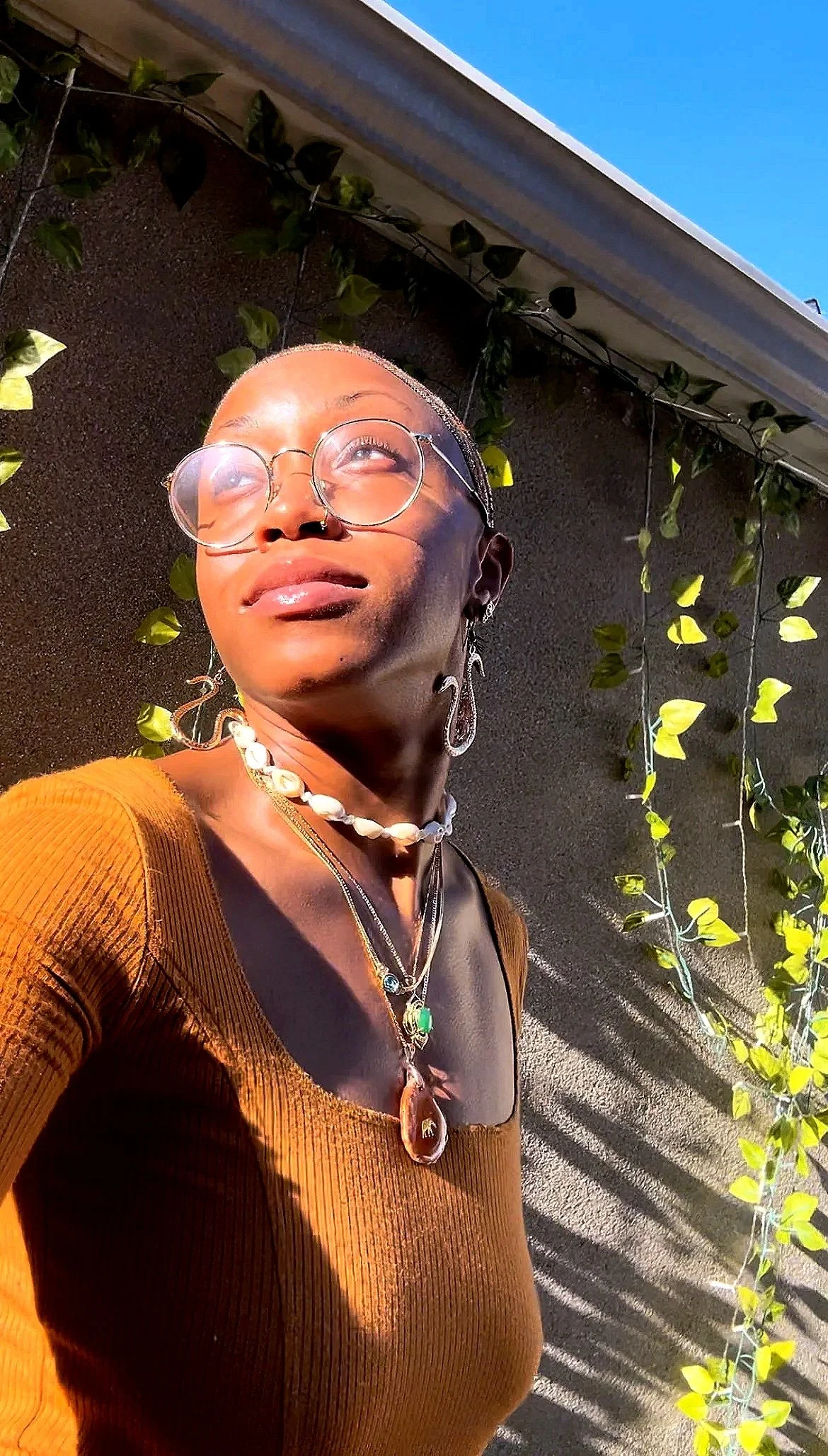 A woman with glasses and jewelry, including earrings and necklaces, taking a selfie outside in sunlight near a wall with decorative greenery.