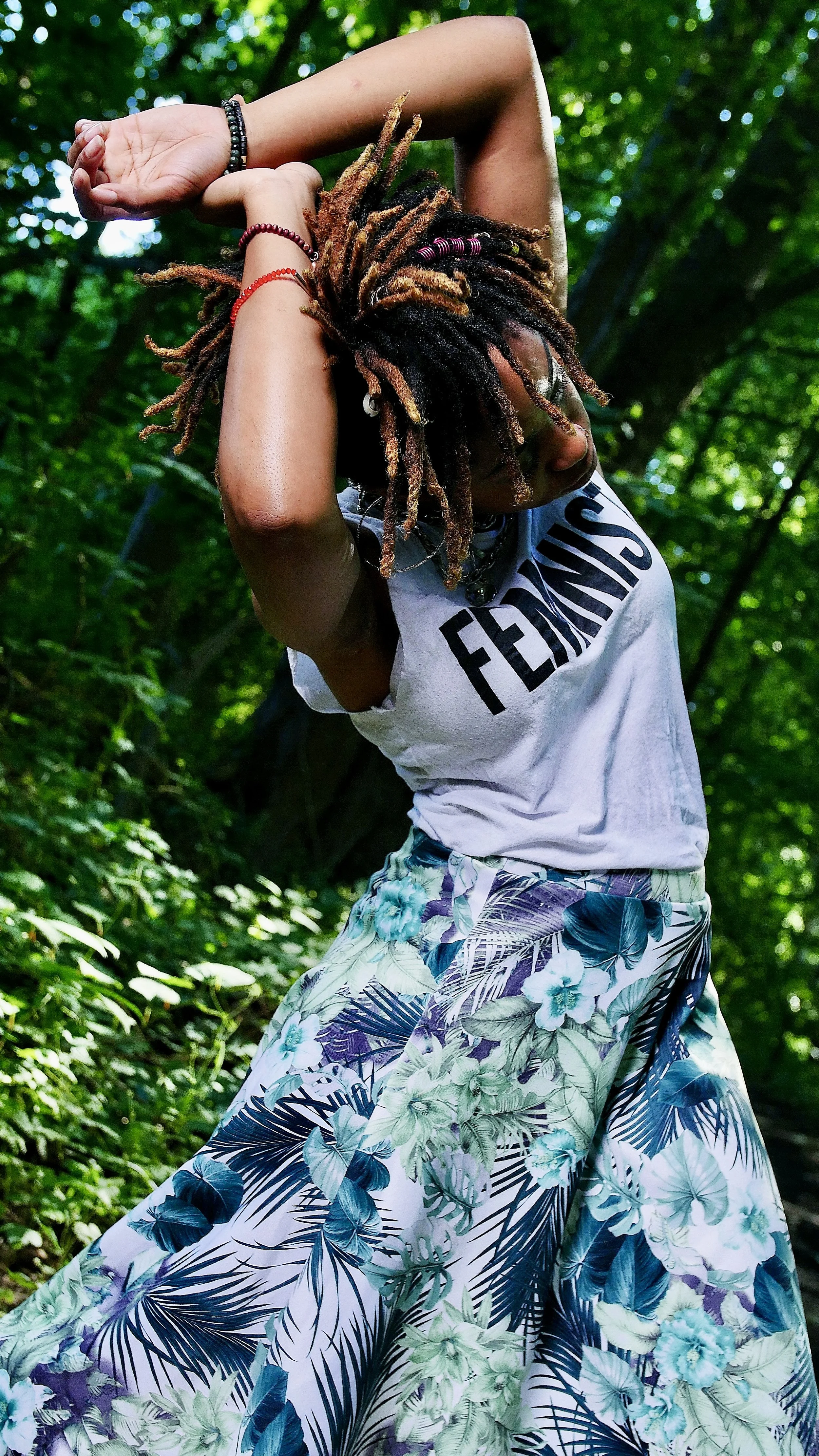 A woman with dreadlocks, wearing a white T-shirt and a floral skirt, standing in a green forest, with her arms raised above her head.