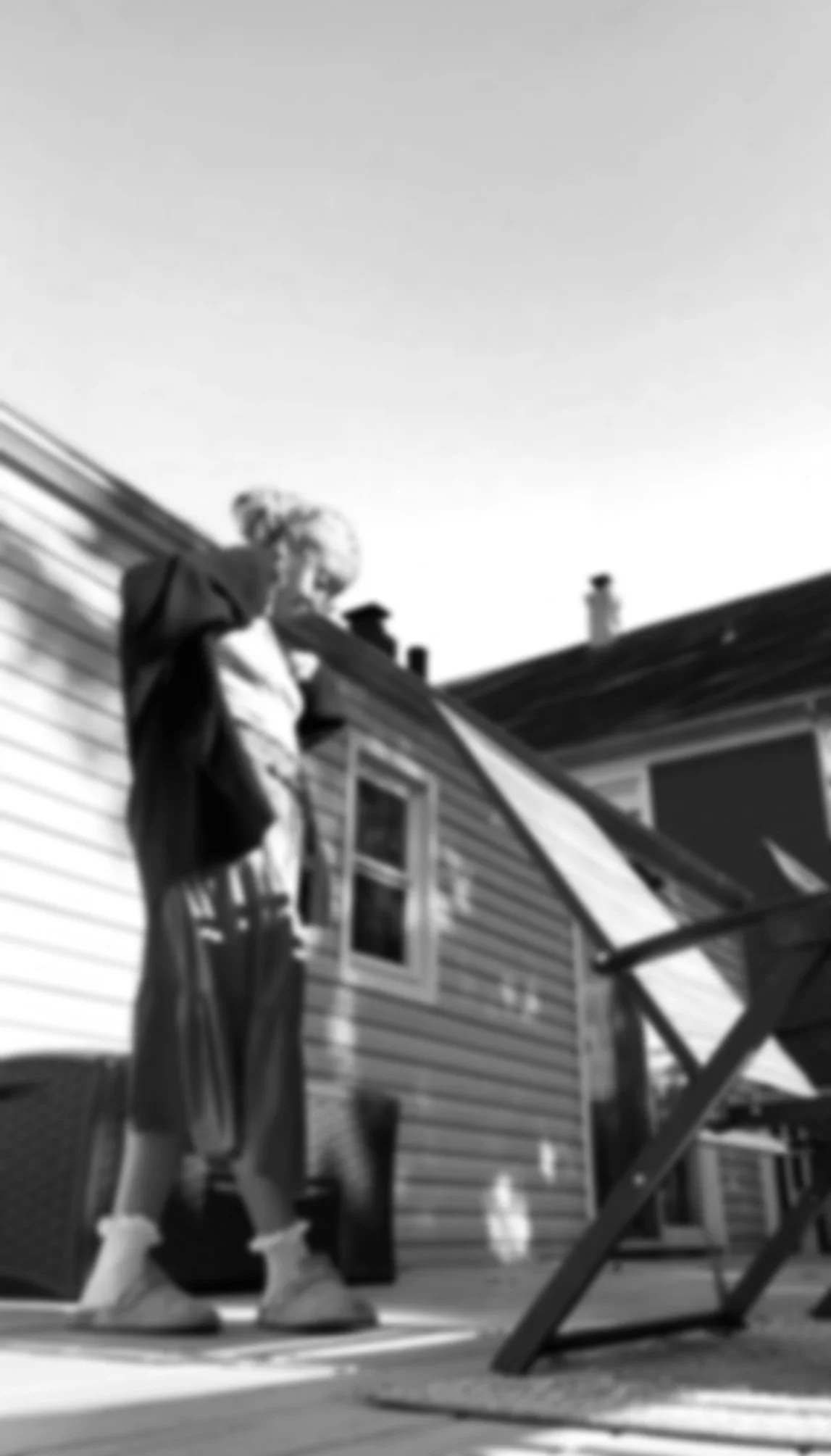 A person in casual clothing standing and leaning on the railing of a house's deck, looking downward, with the back of the house and a clear sky in the background.