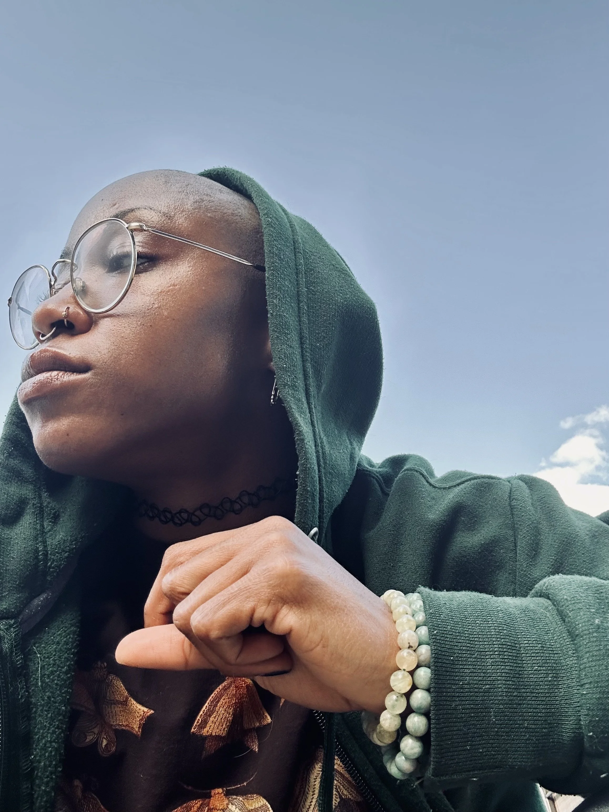 Close-up portrait of a person with glasses, green hoodie, and beaded bracelets, looking to the side with a clear sky in the background.
